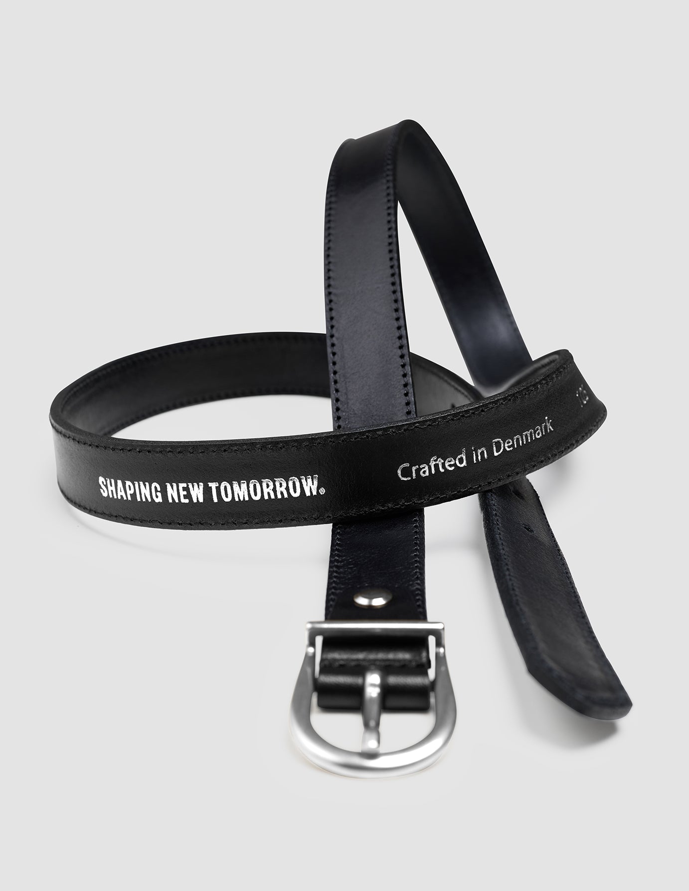 Dünner Damenledergürtel in Silber und Schwarz mit silberner Schnalle, auf dessen Riemen der Schriftzug „SHAPING NEW TOMORROW“ sowie die Prägung „Crafted in Denmark“ zu sehen sind, was das klassische skandinavische Design widerspiegelt.