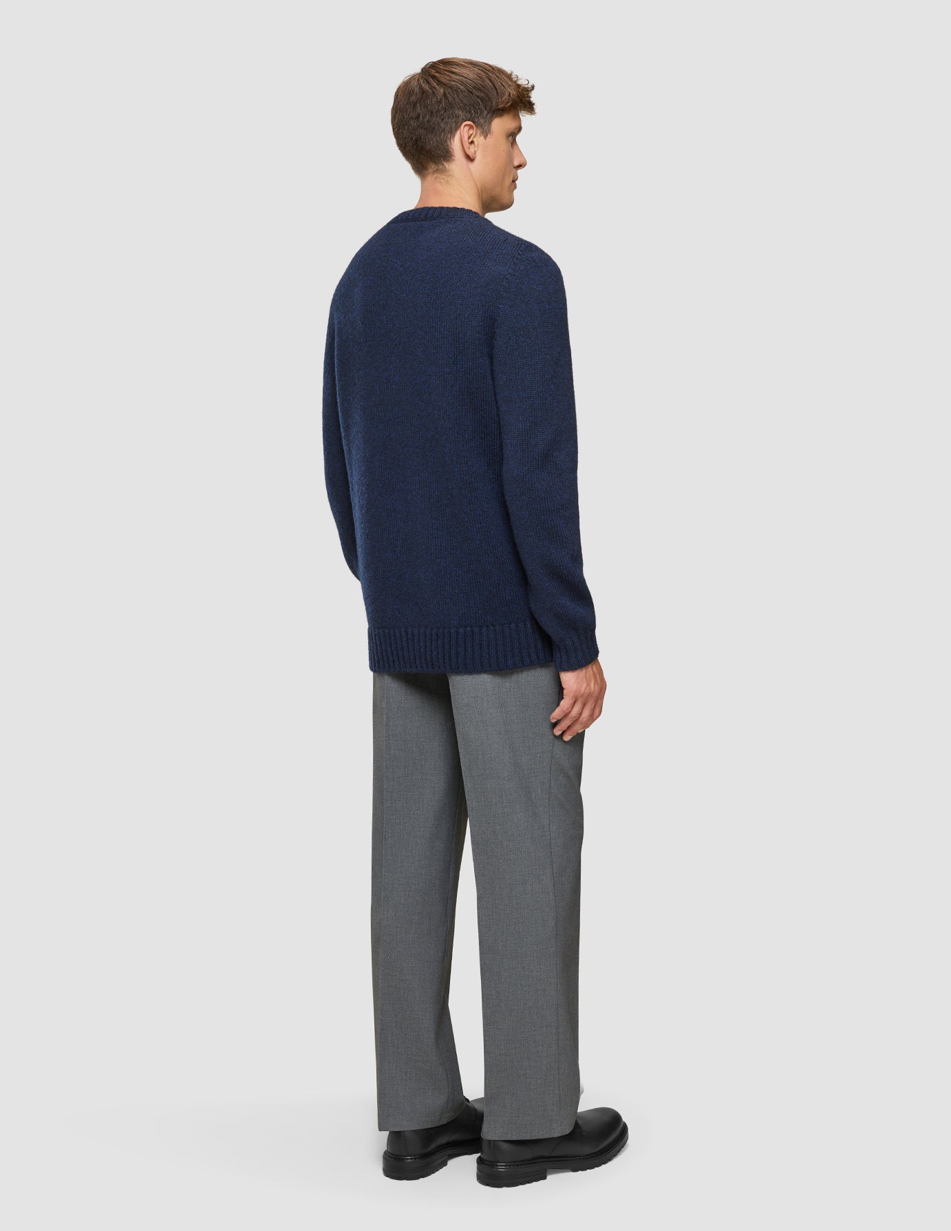 Eine Person, von hinten gesehen, die einen dicken, weichen Wollpullover mit Rundhalsausschnitt in dunklem Marineblau-Melange, eine graue Hose und schwarze Schuhe trägt.