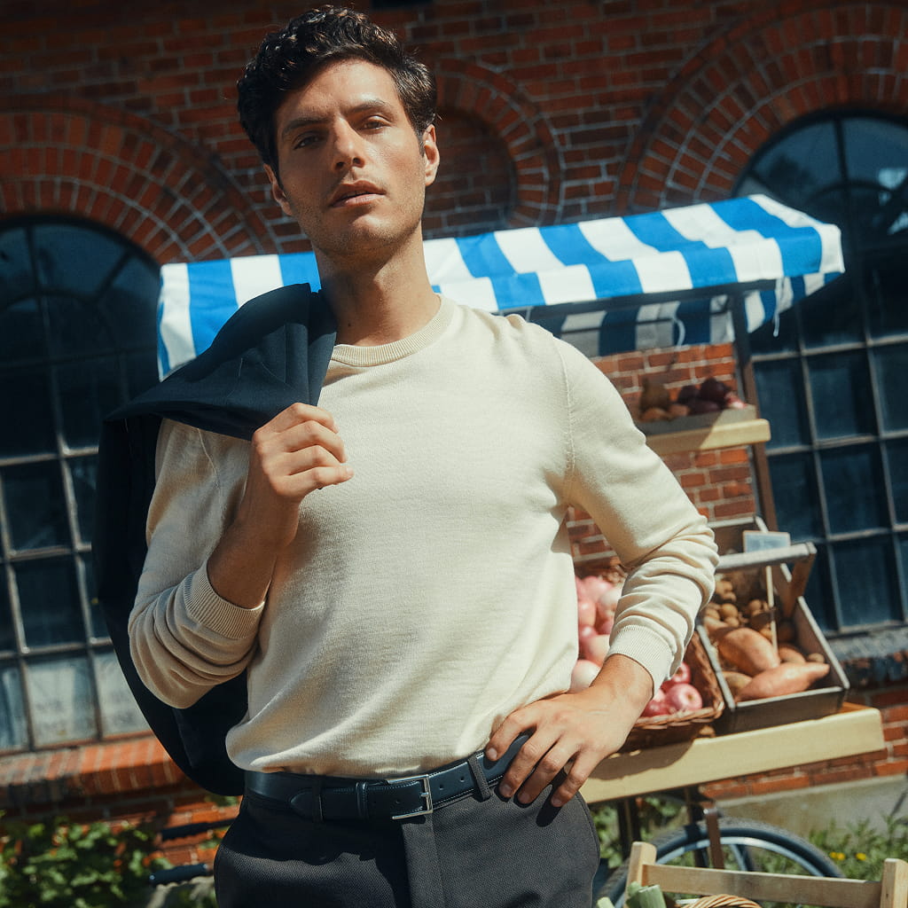 Ein Mann in einem cremefarbenen Pullover, der sich eine Jacke über die Schulter geworfen hat, steht vor einem Obst- und Gemüsestand auf dem Markt.