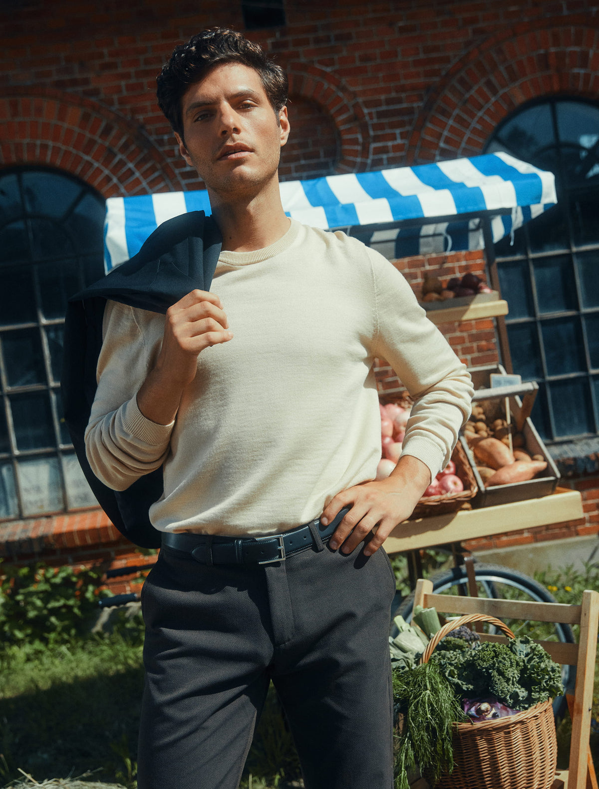 Ein Mann in einem cremefarbenen Pullover und einer dunklen Hose steht vor einem Marktstand, eine Jacke über die Schulter geworfen.