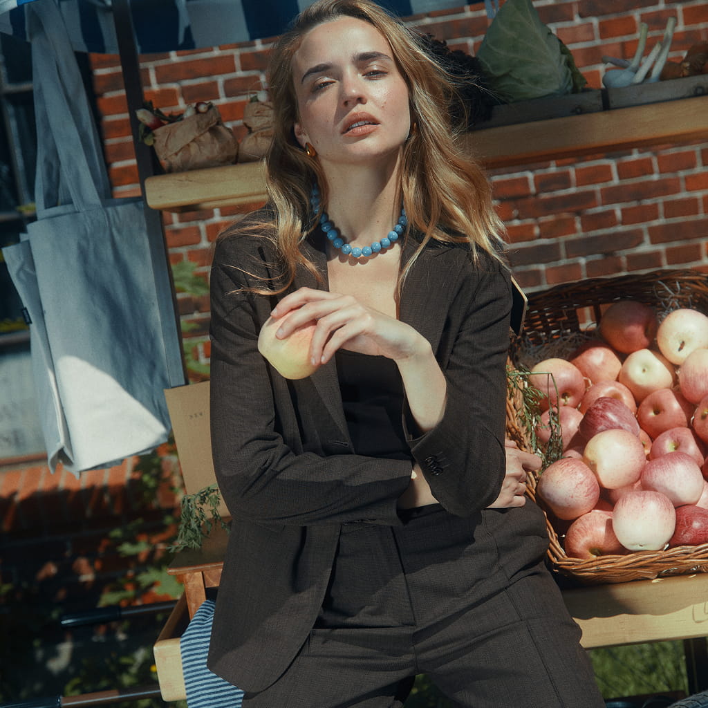 Eine Frau in einem dunklen Kostüm und mit einer blauen Perlenkette hält ein Stück Obst in der Hand, während sie vor einer Kiste mit Äpfeln steht.