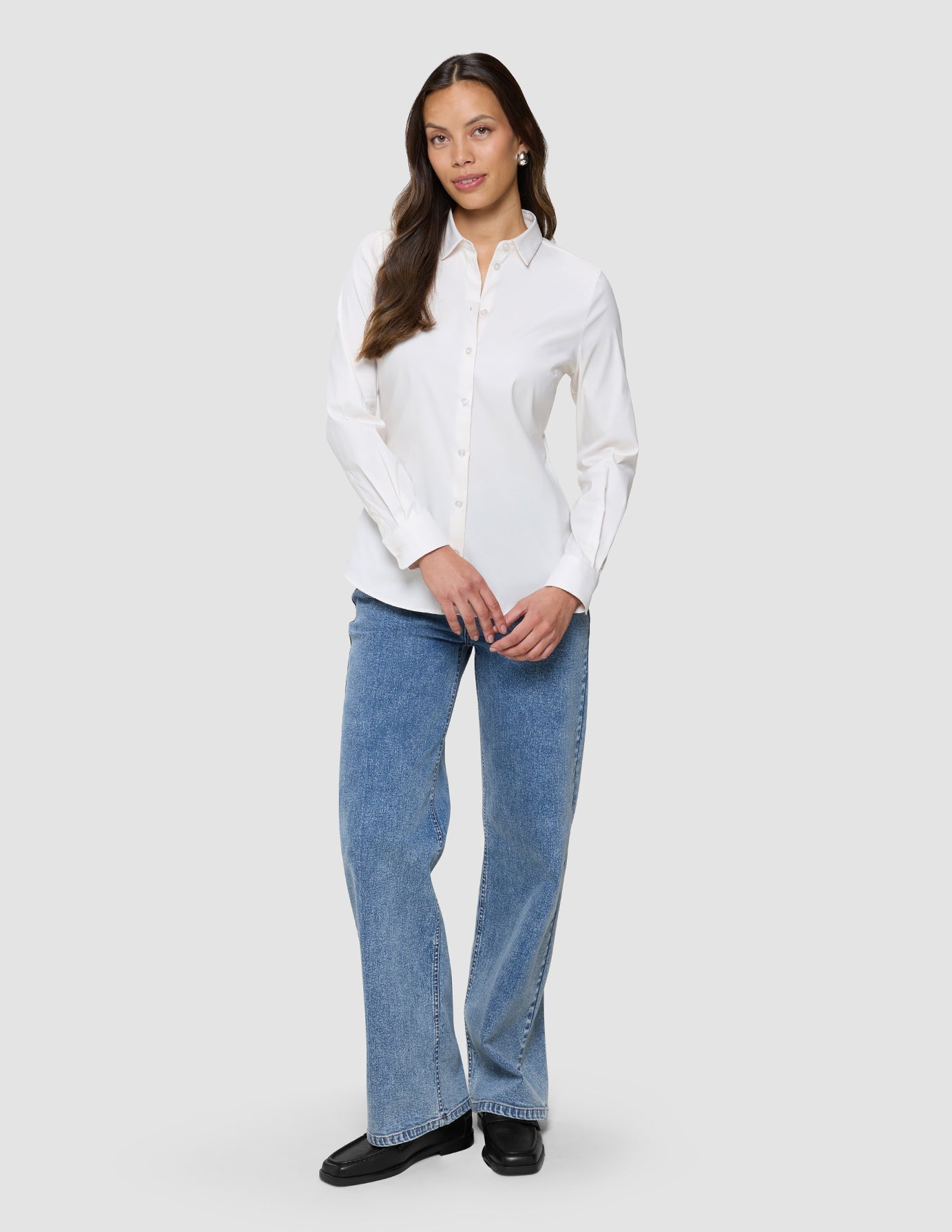 Eine Frau, die ein Damen-Oxford-Hemd White Orchid Slim mit klassischem Kragen trägt, das in eine blaue Jeans gesteckt ist, dazu schwarze Loafer.