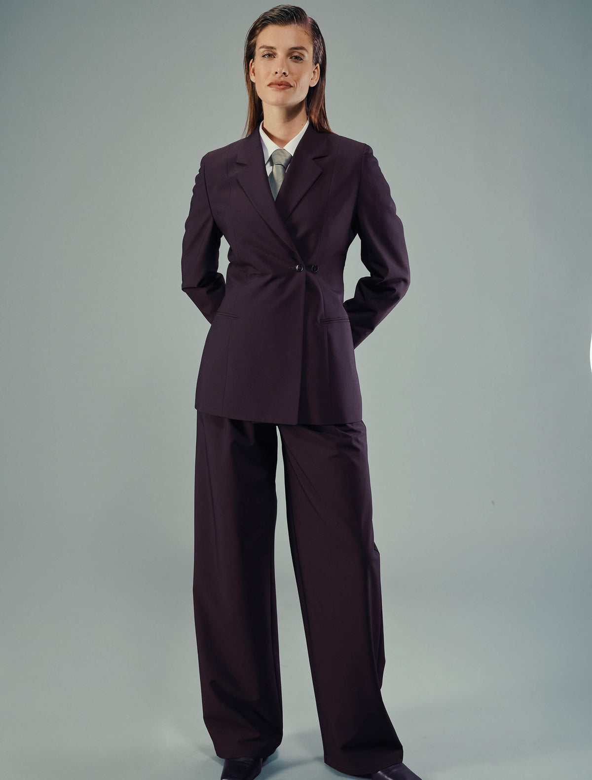 Eine Person, die einen Avira-Blazer für Damen in der Farbe Burgundy Berry und eine dazu passende Hose mit weitem Bein über einem weißen Hemd und einer Krawatte trägt, was ein minimalistisches skandinavisches Design widerspiegelt.
