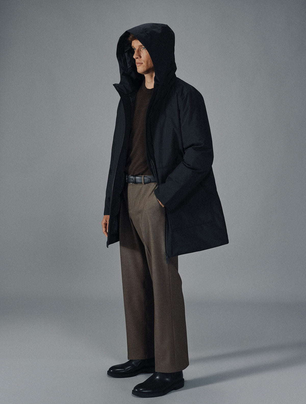 Ein Mann, der eine schwarze Utility-Jacke, einen dunkelbraunen Pullover, eine braune Hose und schwarze Schuhe trägt.