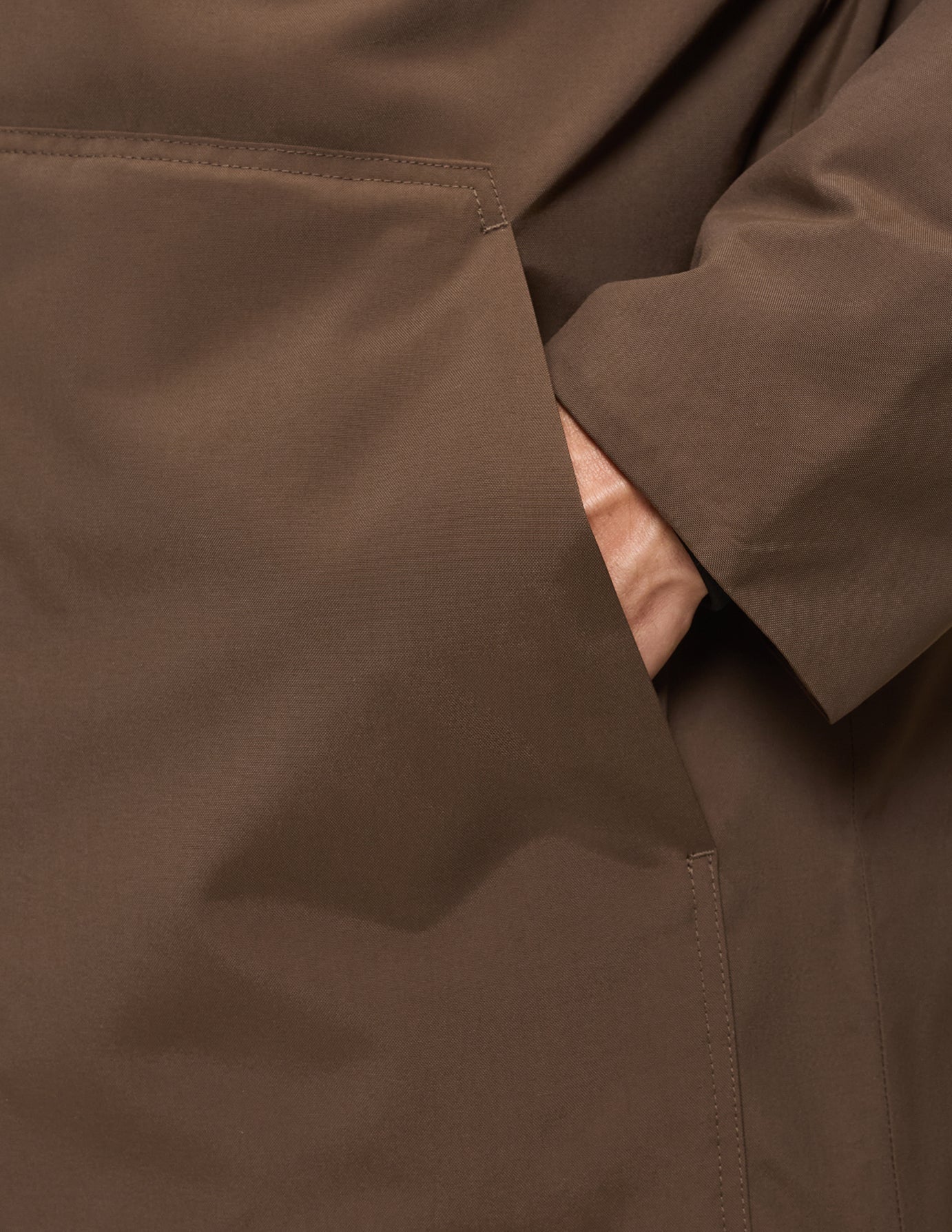 Eine Hand, die in der Seitentasche einer winddichten Utility-Jacke in Sattelbraun ruht.