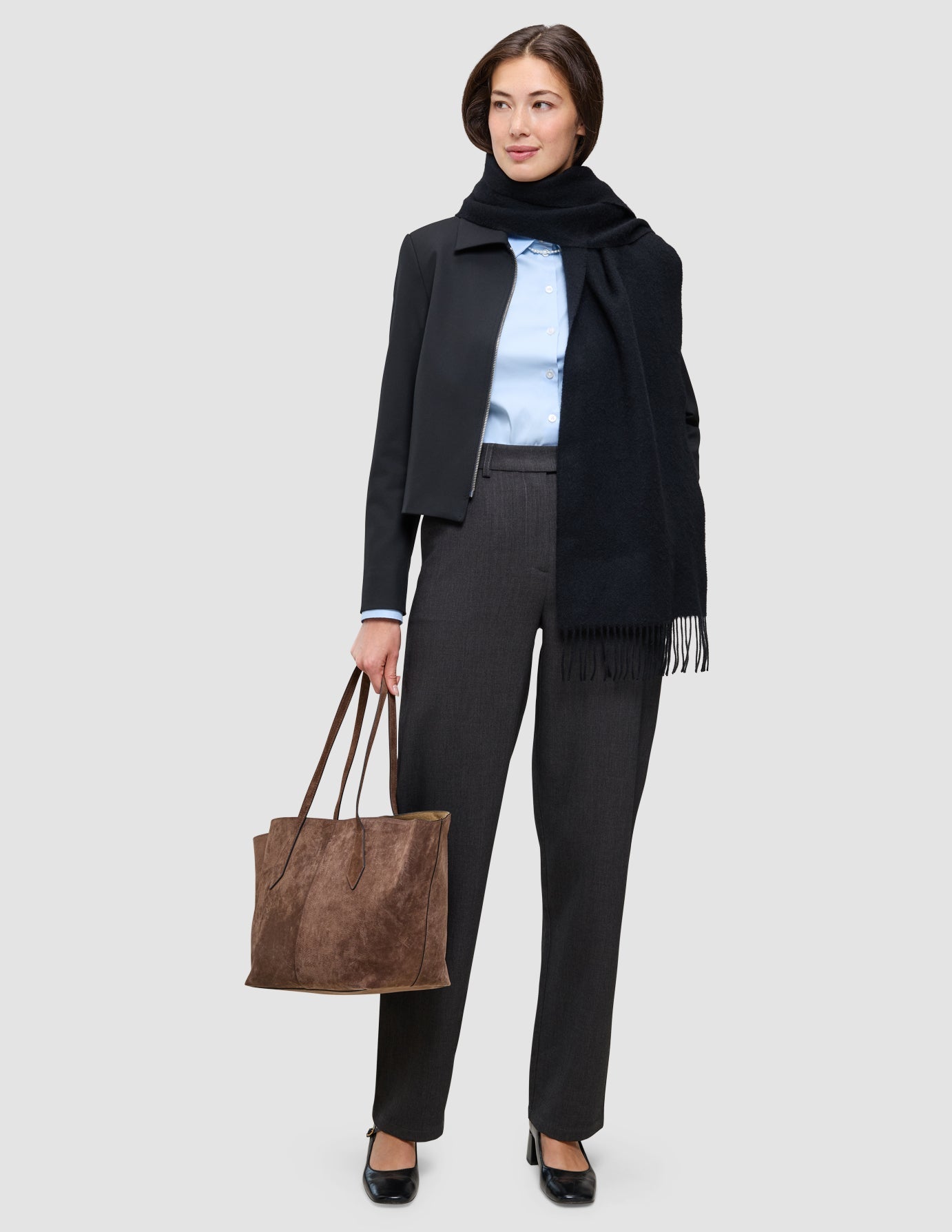 Eine Person, die ein hellblaues Oxford-Hemd, eine dunkle Hose und einen schwarzen Schal trägt – eine einfache Aufwertung des Büro-Looks – und eine große braune Tragetasche bei sich hat.
