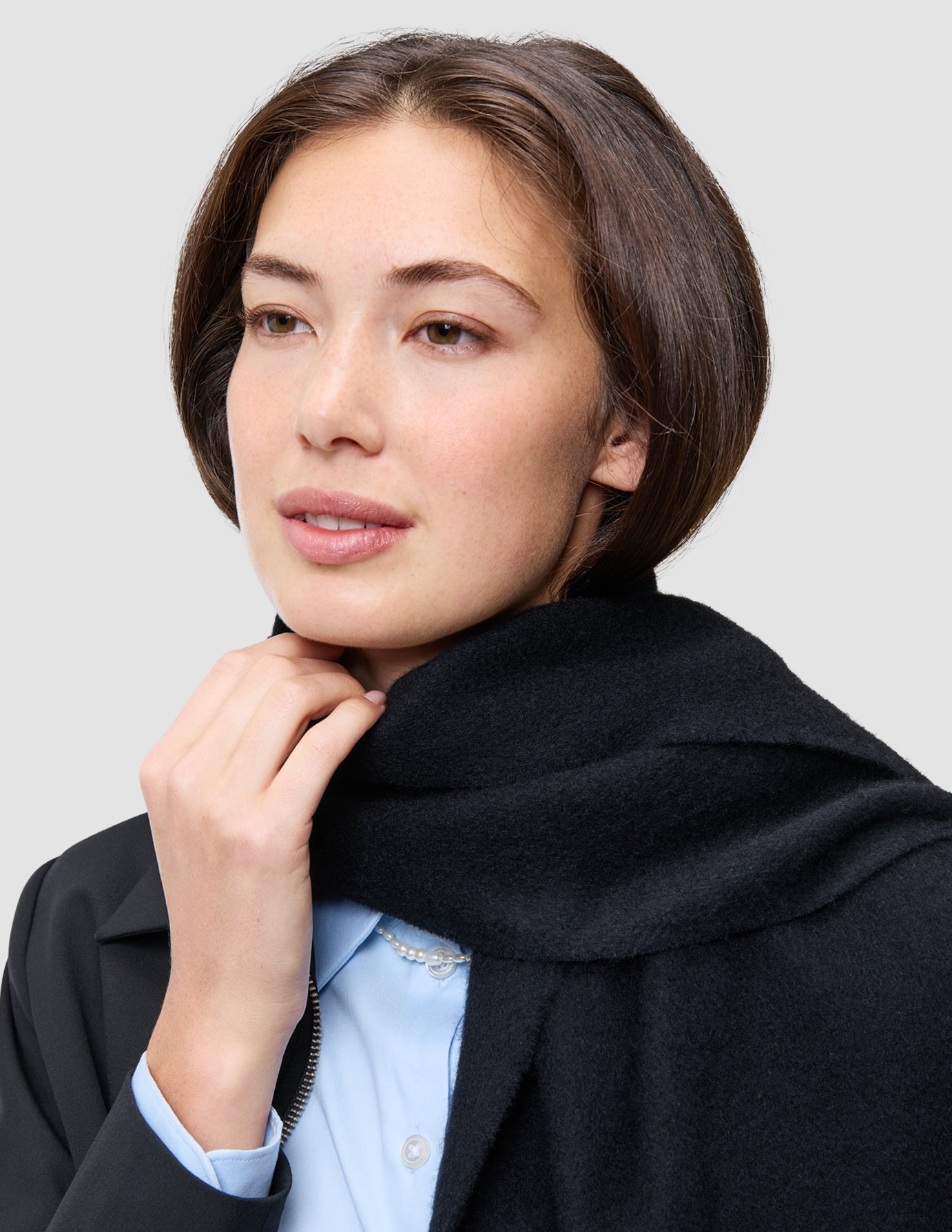 Eine Frau, die eine schwarze Essential Zip Jacket von An easy office upgrade über einem hellblauen Oxford-Hemd und einem schwarzen Schal trägt.