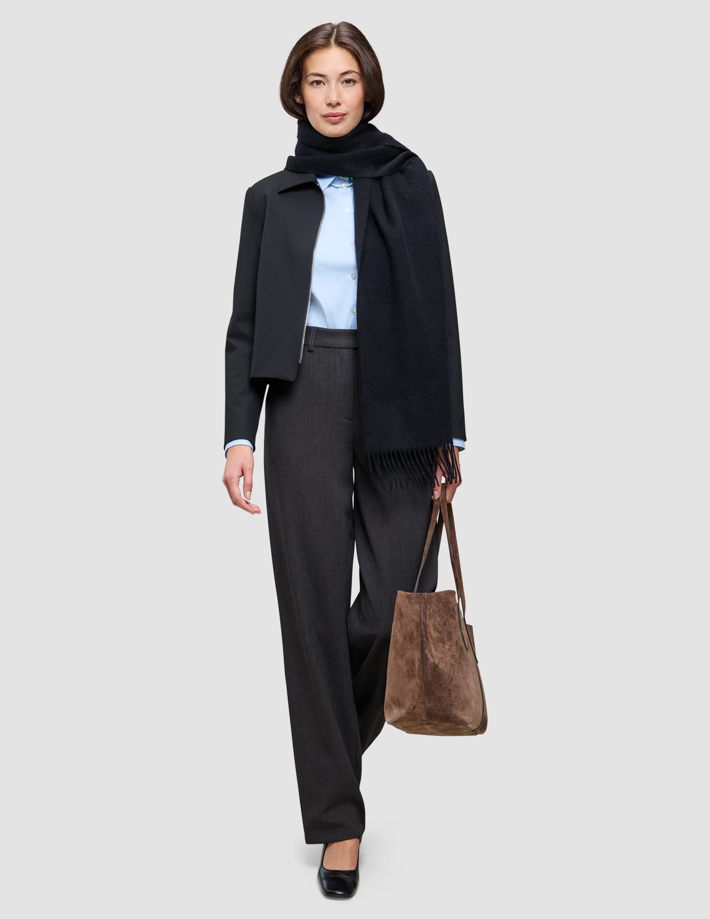 Eine Frau trägt eine lässige Jacke, die ihr Outfit für das Büro aufwertet, über einem hellblauen Oxford-Hemd und einer anthrazitfarbenen Hose, dazu einen schwarzen Schal und eine braune Tragetasche.
