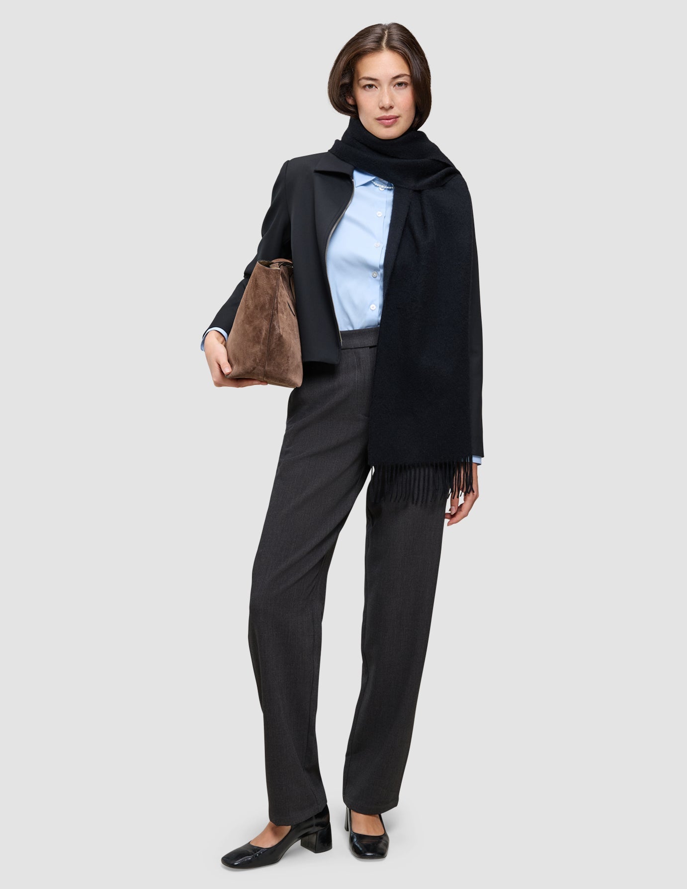 Eine Frau, die ein hellblaues An easy office upgrade-Outfit, einen schwarzen Blazer und eine Go2-Hose trägt, eine braune Handtasche in der Hand hält und einen schwarzen Schal trägt.