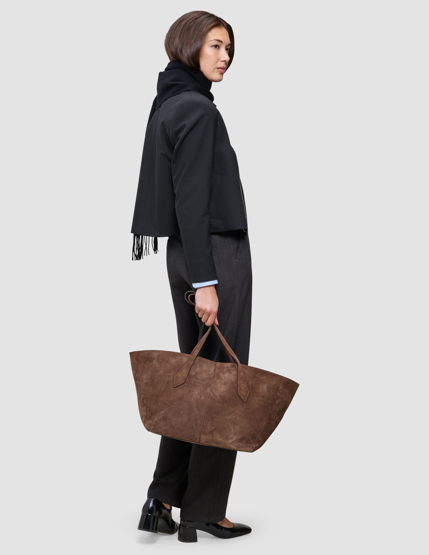Eine Frau, die eine An easy office upgrade-Jacke mit Reißverschluss und eine Hose trägt, hält eine große braune Wildleder-Tragetasche in der Hand.