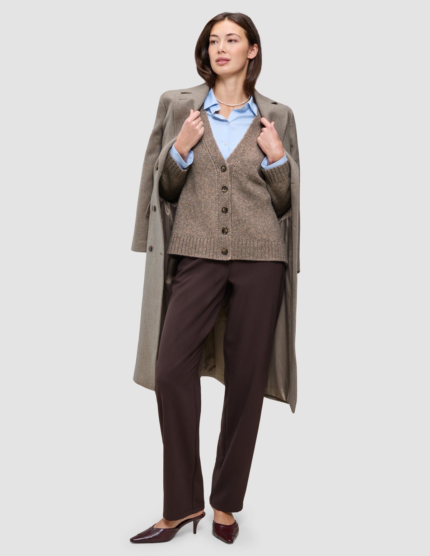 Eine Frau in Winterarbeitskleidung trägt eine braune Strickjacke über einem hellblauen Oxford-Hemd und einer dunklen Go2-Hose, über die Schultern hat sie einen langen Wollmantel geworfen.