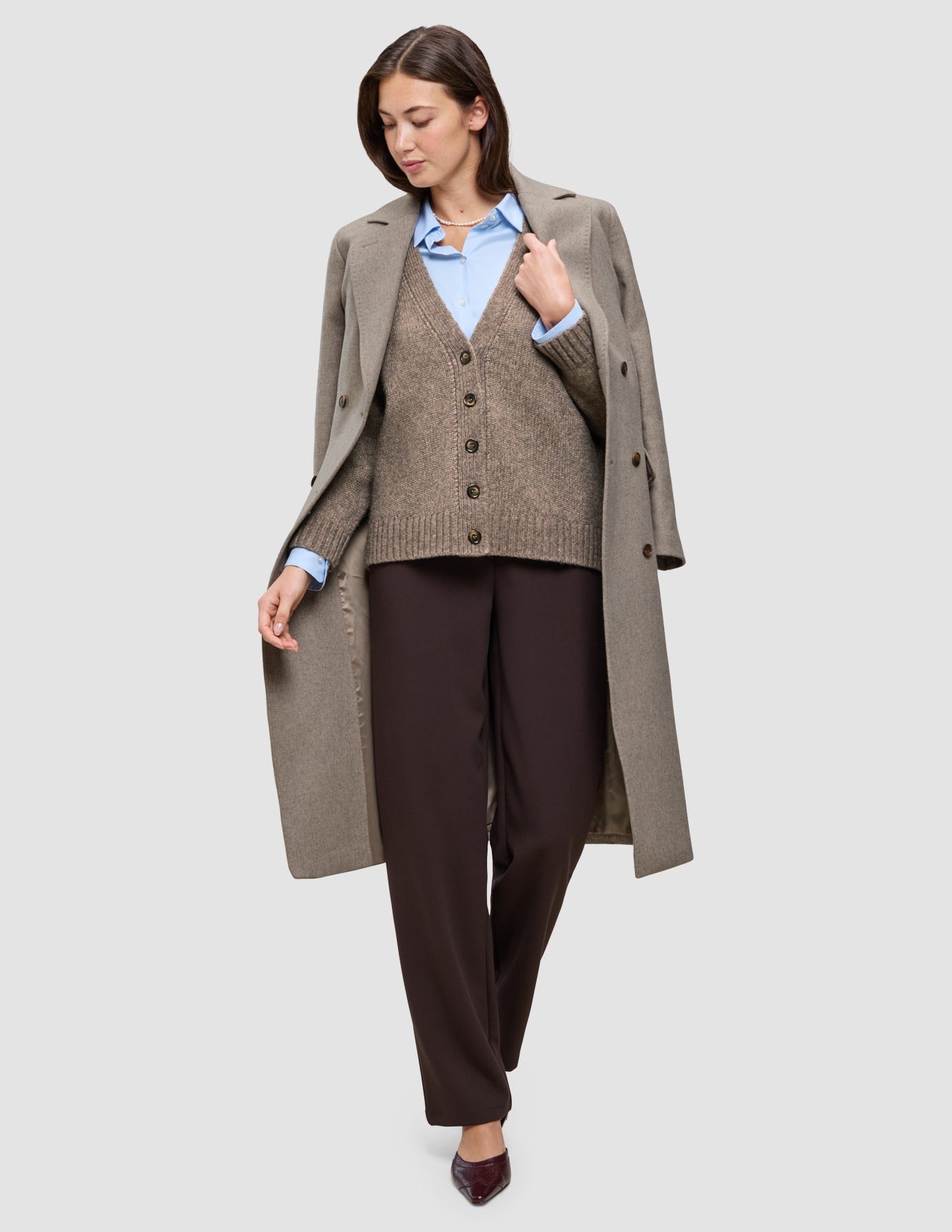 Eine Person, die Winterarbeitskleidung trägt, bestehend aus einem langen Wollmantel über einer braunen Strickjacke, einem hellblauen Oxford-Hemd und einer dunklen Hose.