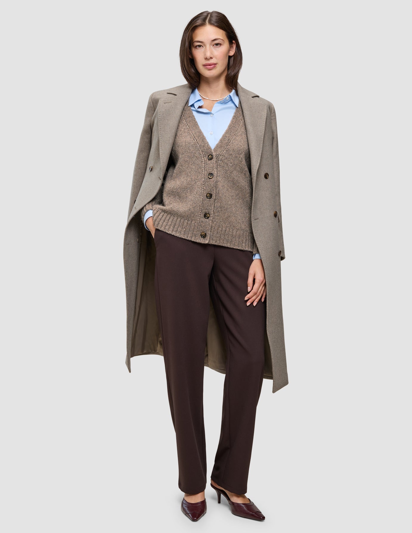 Eine Frau, die einen hellbraunen Wollmantel über einer braunen Strickjacke mit Knöpfen, einem hellblauen Oxford-Hemd und einer Winterarbeitshose trägt.