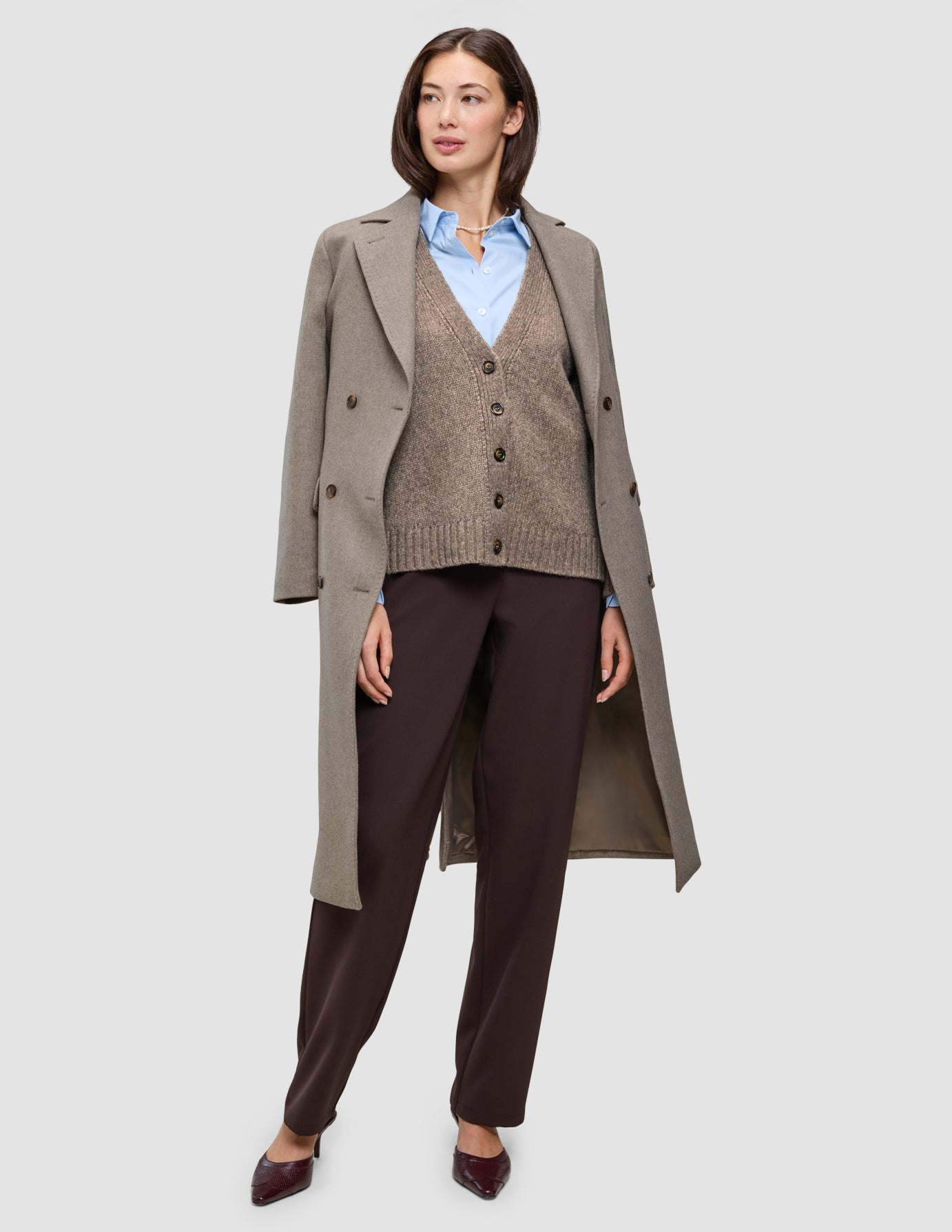Eine Frau trägt Winterarbeitskleidung, bestehend aus einem langen Wollmantel über einem hellblauen Oxford-Hemd, einer braunen Strickjacke mit V-Ausschnitt und einer dunkelbraunen Go2-Hose.