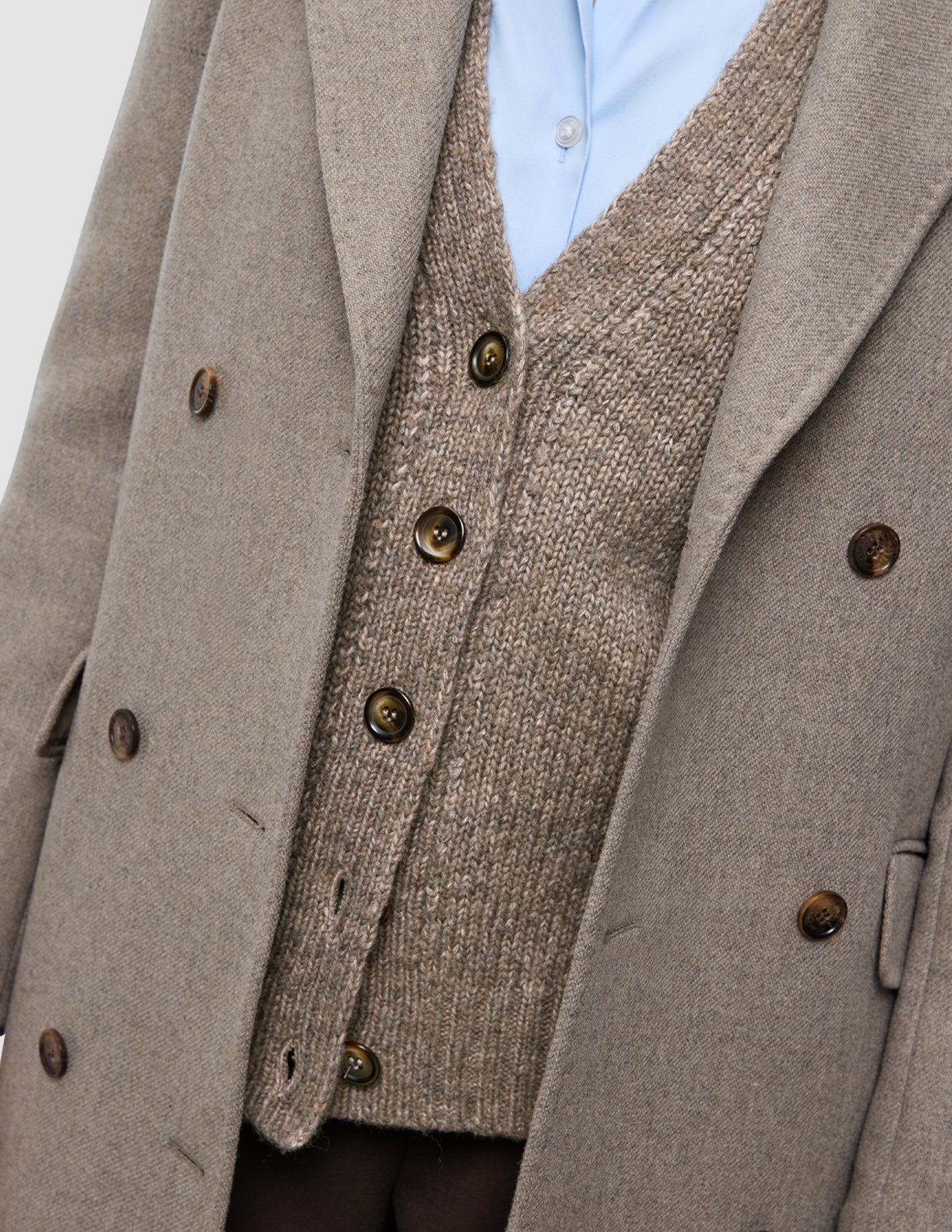 Arbeitskleidung für kaltes Wetter, bestehend aus einem hellbraunen, zweireihigen Wollmantel, der über einem hellblauen Oxford-Hemd und einer braunen, geknöpften Strickjacke getragen wird.