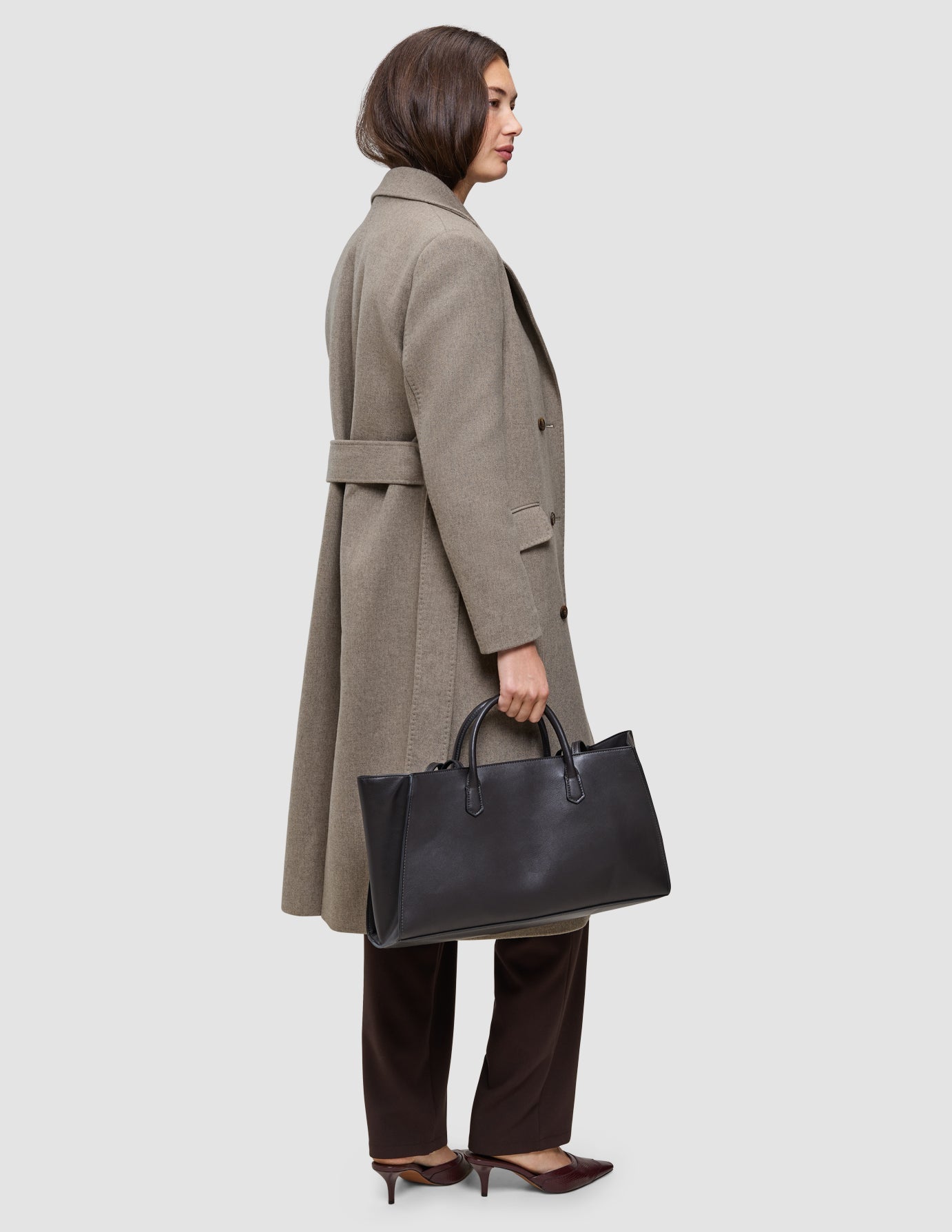 Eine Person, die einen langen, mit einem Gürtel geschnürten taupefarbenen Wollmantel über einer Arbeitshose für kaltes Wetter trägt und eine große schwarze Handtasche bei sich hat.