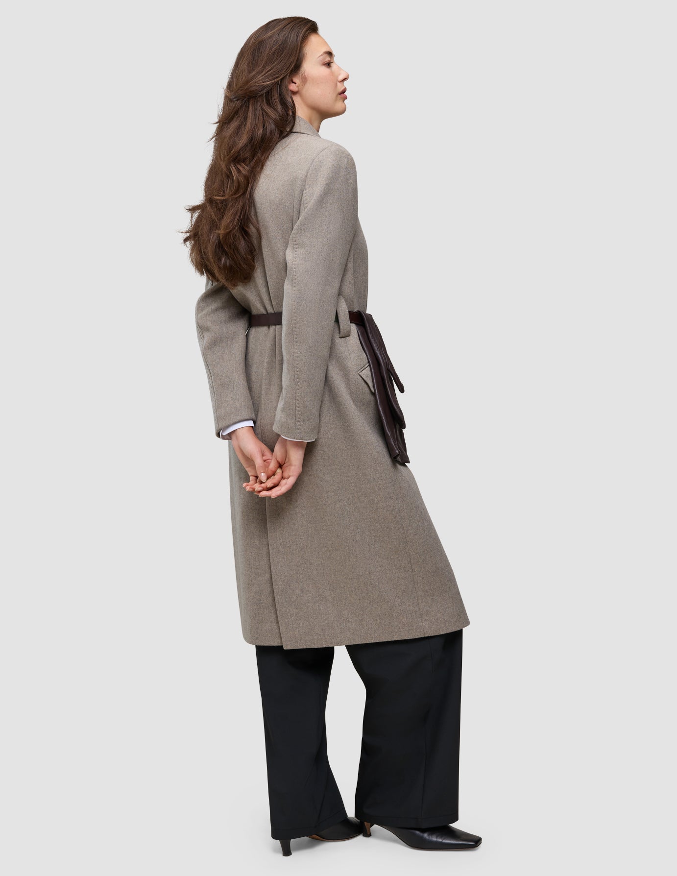 Eine Frau, die einen winterlichen, eleganten, knielangen, hellbraunen Wollmantel mit einem dunkelbraunen Gürtel und einer Avira-Hose trägt.