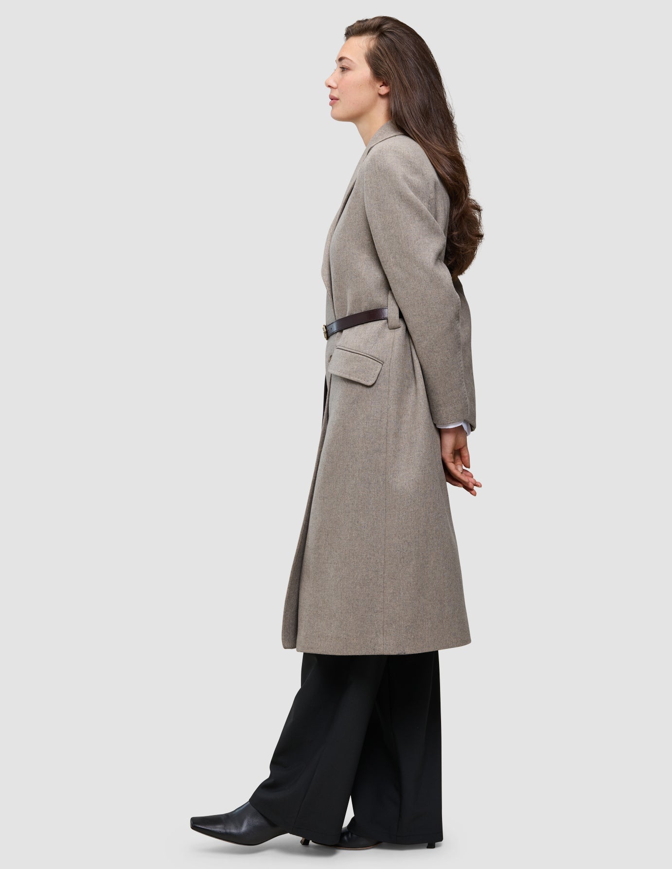 Eine Person, die einen winterlichen, eleganten Wollmantel trägt, der mit einem dunklen, schmalen Gürtel tailliert ist, dazu eine schwarze Avira-Hose und schwarze Stiefel mit spitzer Zehenpartie.