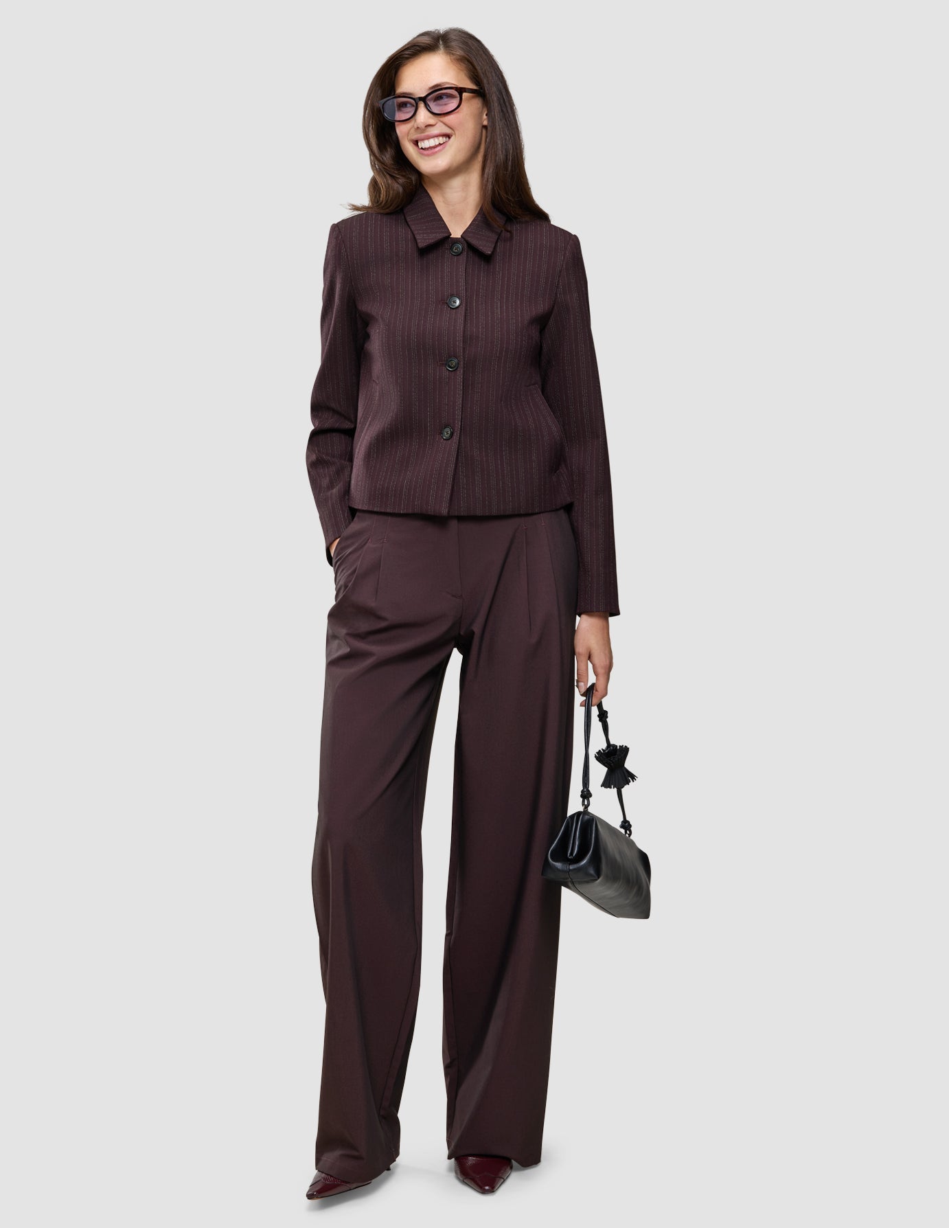 Eine Frau, die eine dunkelburgunderrote Nadelstreifenjacke und eine dazu passende Hose mit weitem Bein aus der Kollektion Burgundy from top to toe trägt und eine kleine schwarze Handtasche in der Hand hält.