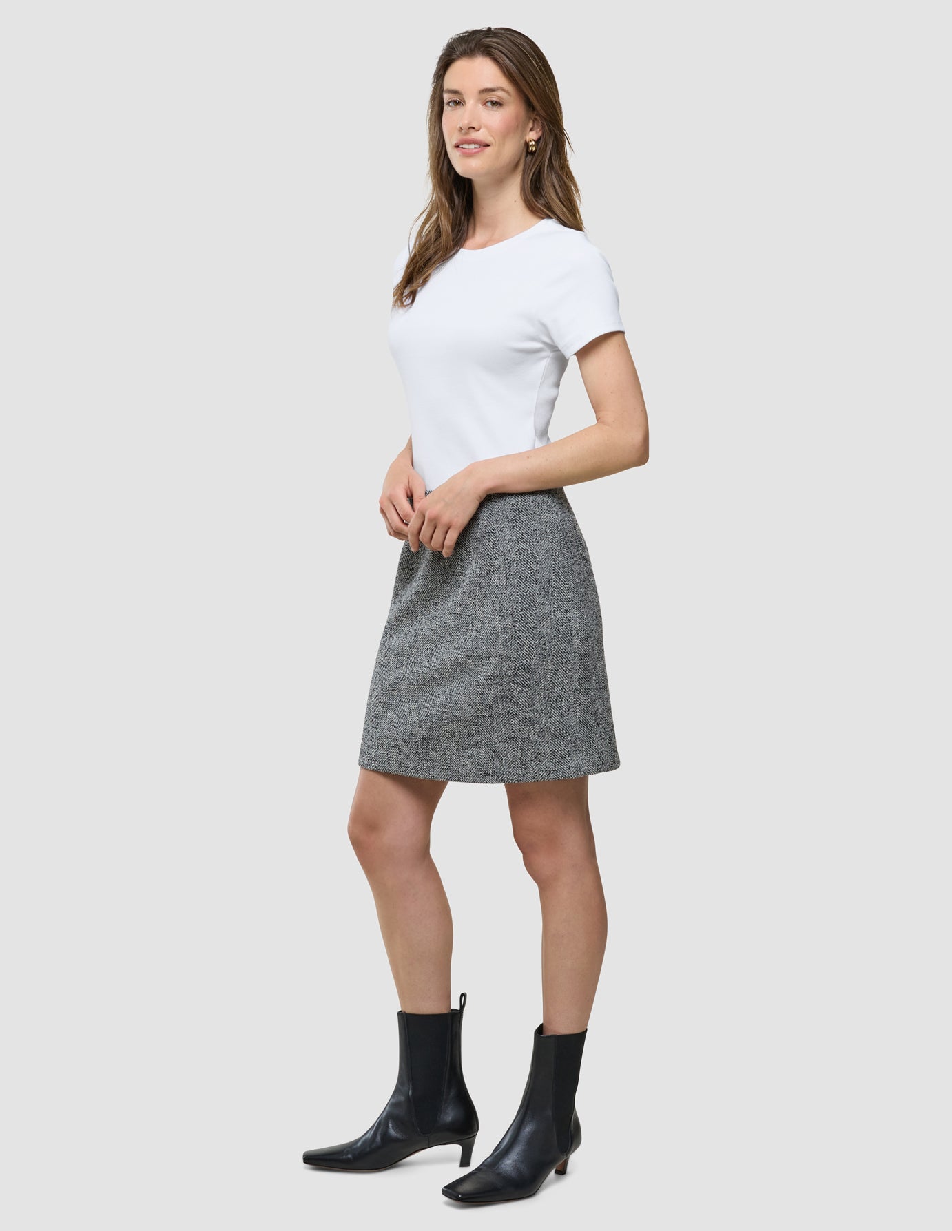 Eine Frau, die ein weißes T-Shirt, einen kurzen „Ashryn“-Rock von Domino mit Fischgrätenmuster und schwarze Stiefeletten trägt, steht vor einem einfarbigen Hintergrund.
