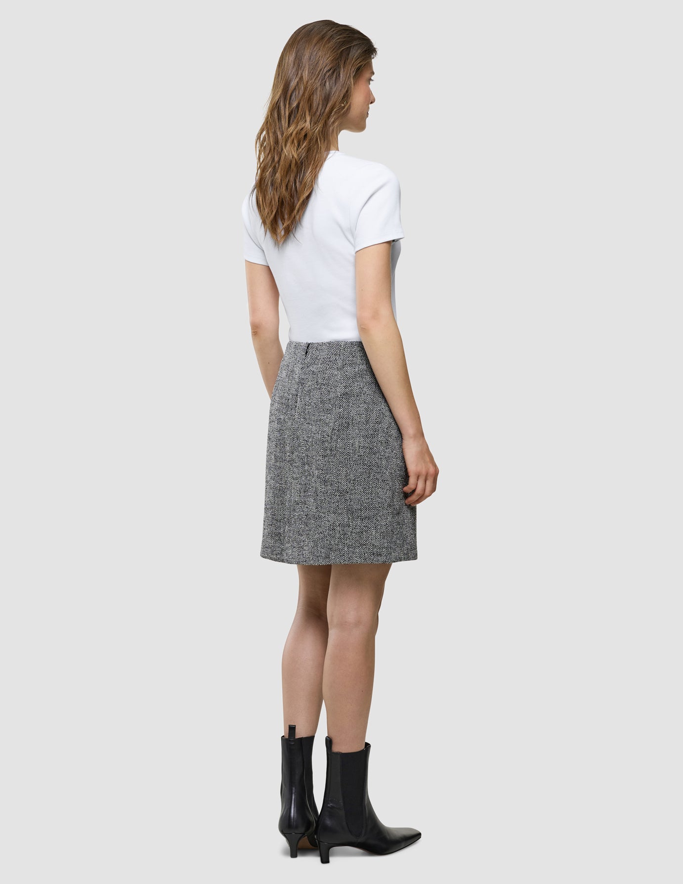 Eine Frau, von hinten gesehen, die ein weißes T-Shirt, einen „Women Ashryn Short Skirt Domino Herringbone“-Rock und schwarze Stiefeletten trägt.