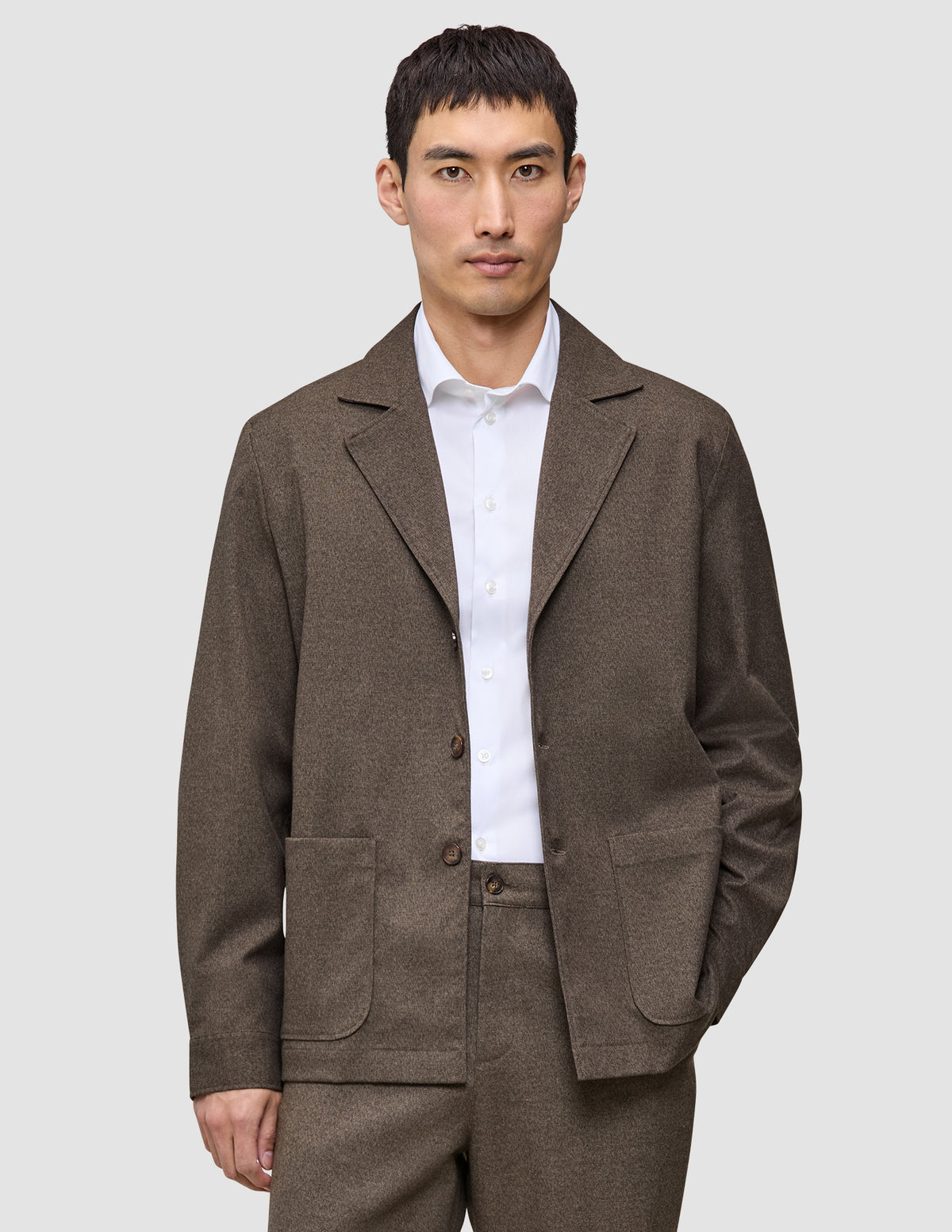 Ein Mann, der ein Atelier Overshirt aus dunkelbeigem Broken-Twill über einem weißen Hemd mit Kragen trägt.