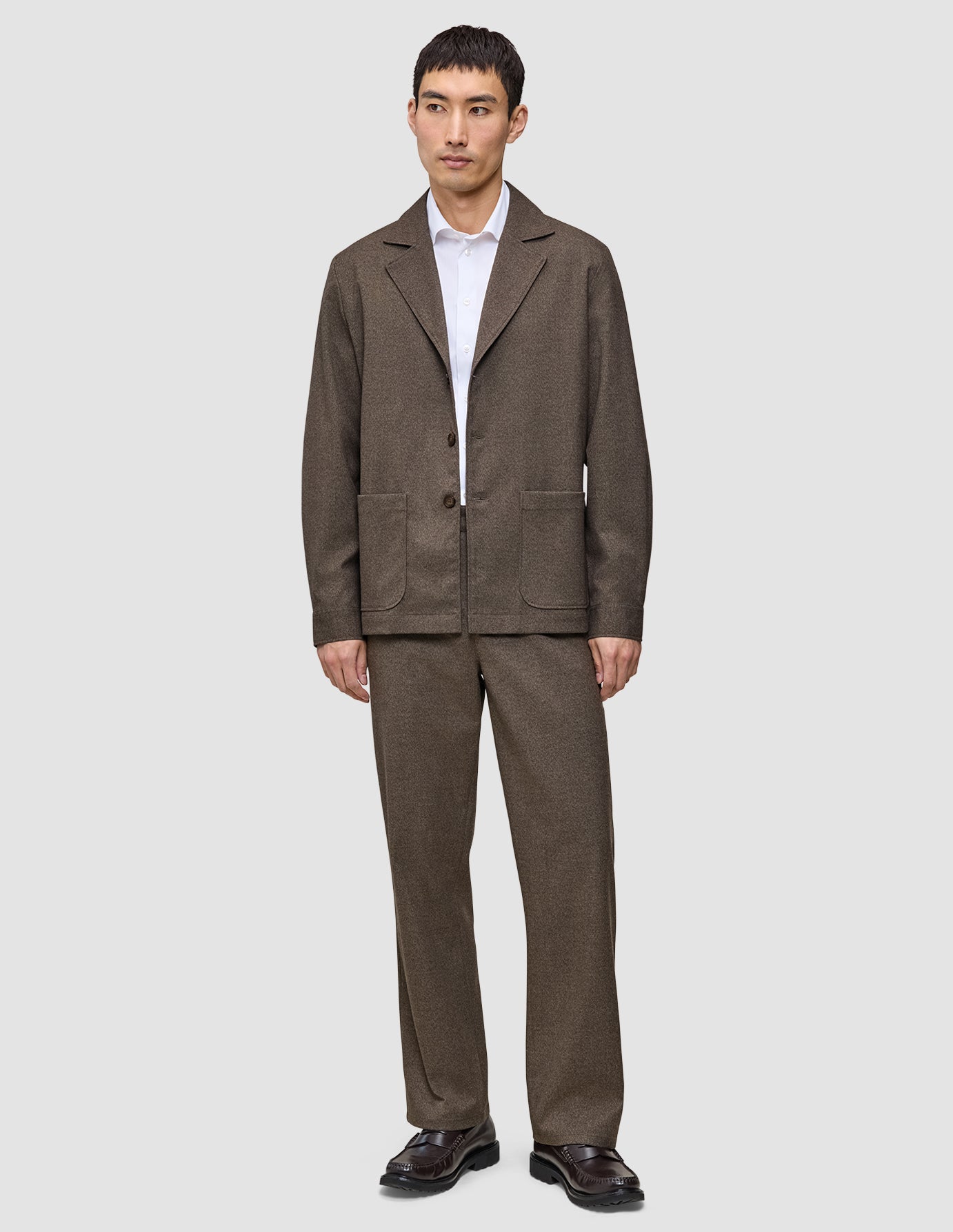 Ein Mann trägt ein Atelier Overshirt Dark Beige aus gebrochenem Twill mit einer dazu passenden Hose über einem weißen Hemd mit Kragen, ergänzt durch dunkle Loafer – ein Beispiel für skandinavisches Design.