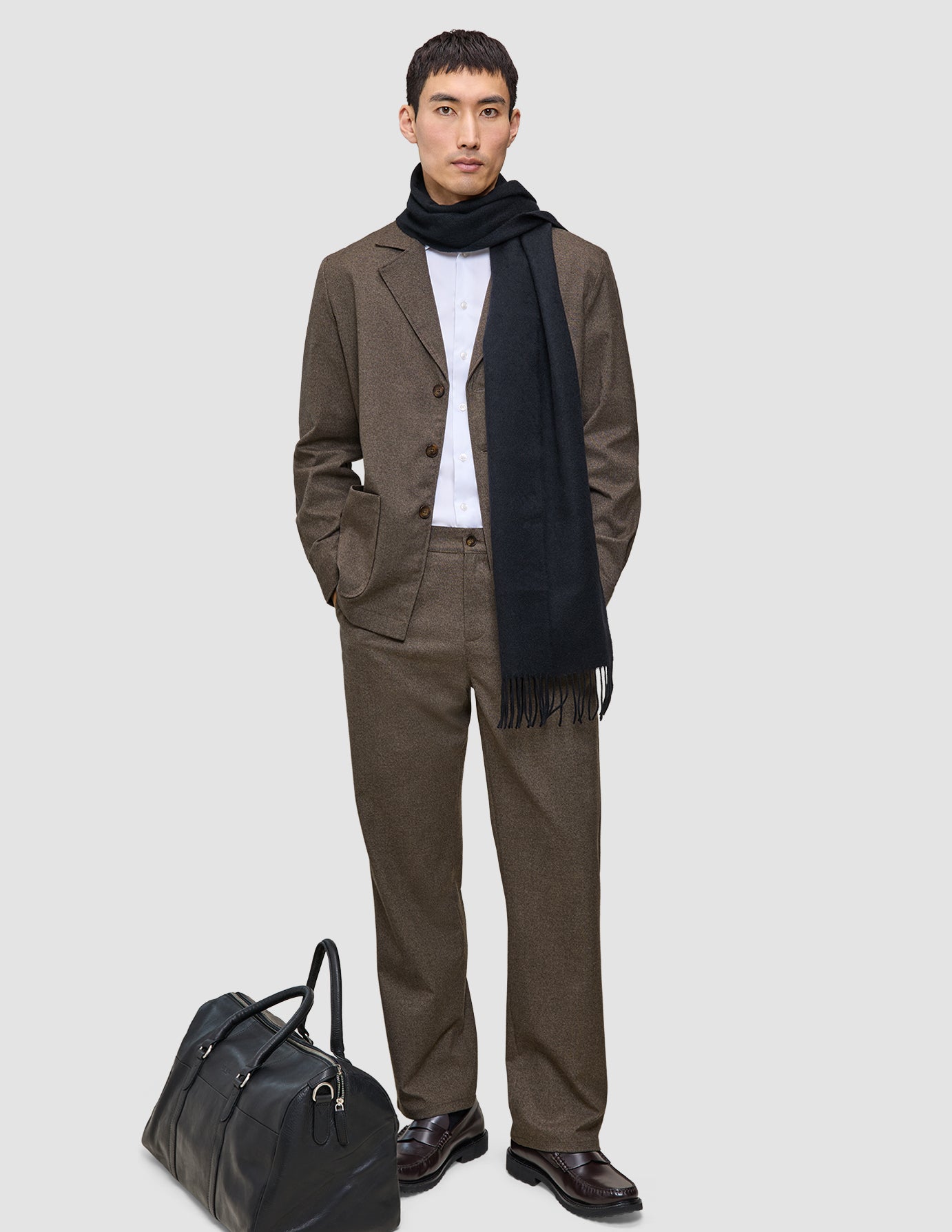 Ein Mann trägt ein beiges Atelier Overshirt Dark Beige über einem weißen Hemd und einem dunklen Schal; zu seinen Füßen steht eine schwarze Reisetasche.