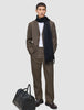 Ein Mann trägt ein beiges Atelier Overshirt Dark Beige über einem weißen Hemd und einem dunklen Schal; zu seinen Füßen steht eine schwarze Reisetasche.