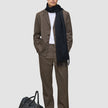 Ein Mann trägt ein beiges Atelier Overshirt Dark Beige über einem weißen Hemd und einem dunklen Schal; zu seinen Füßen steht eine schwarze Reisetasche.