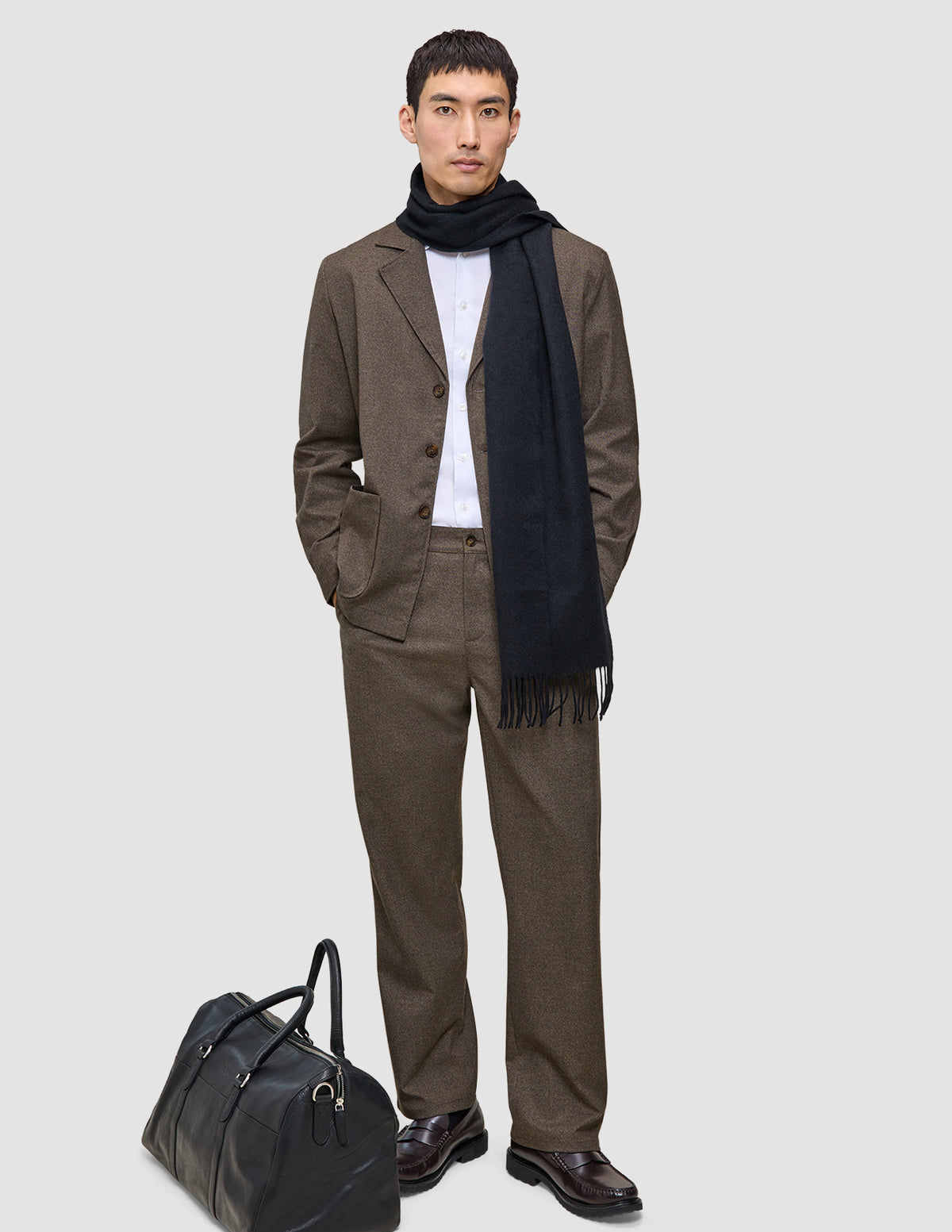 Ein Mann trägt ein beiges Atelier Overshirt Dark Beige über einem weißen Hemd und einem dunklen Schal; zu seinen Füßen steht eine schwarze Reisetasche.