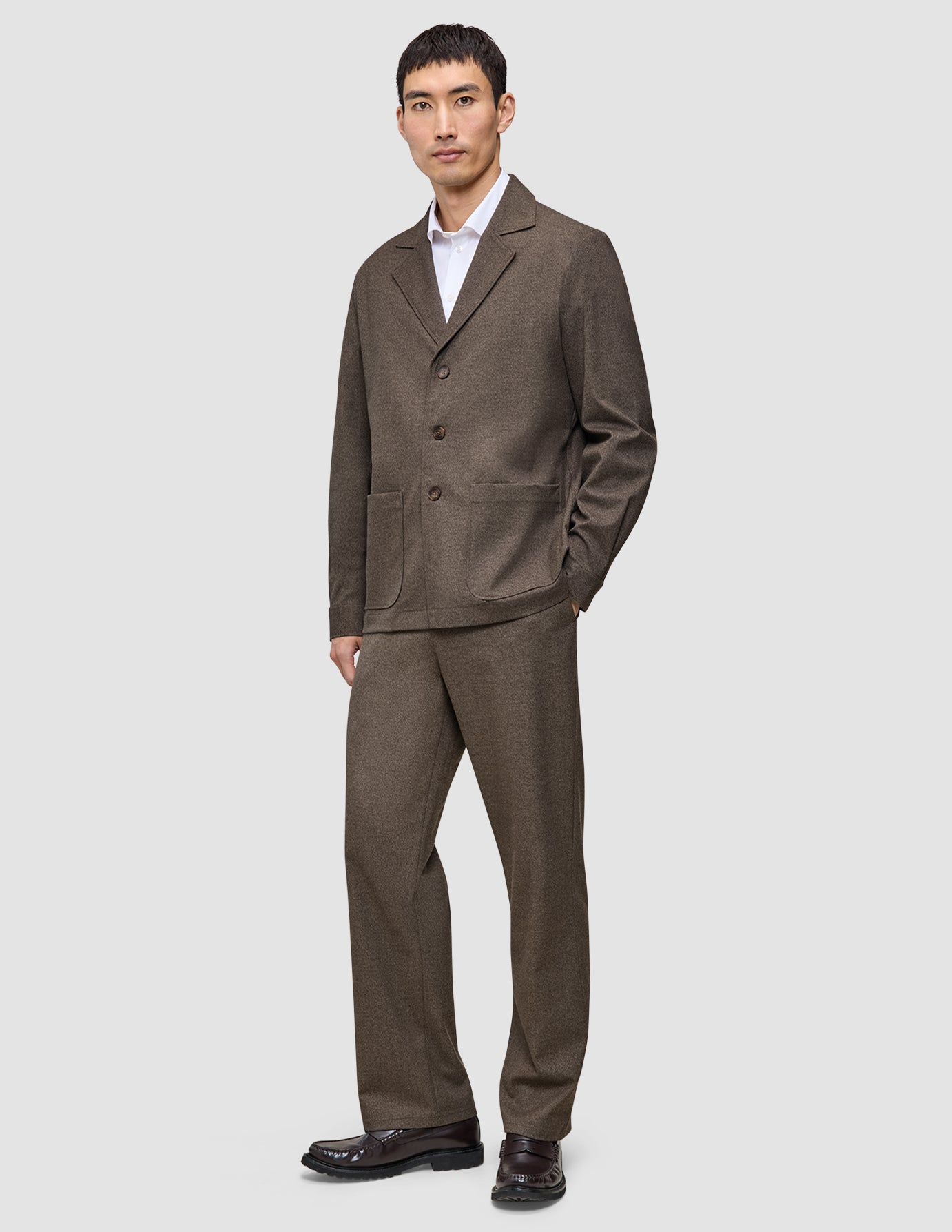 Ein Mann, der ein dunkelbeiges Atelier Overshirt aus gebrochenem Twill über einem weißen Hemd mit Kragen trägt, dazu eine passende Hose und dunkle Loafer im klaren, skandinavischen Design.