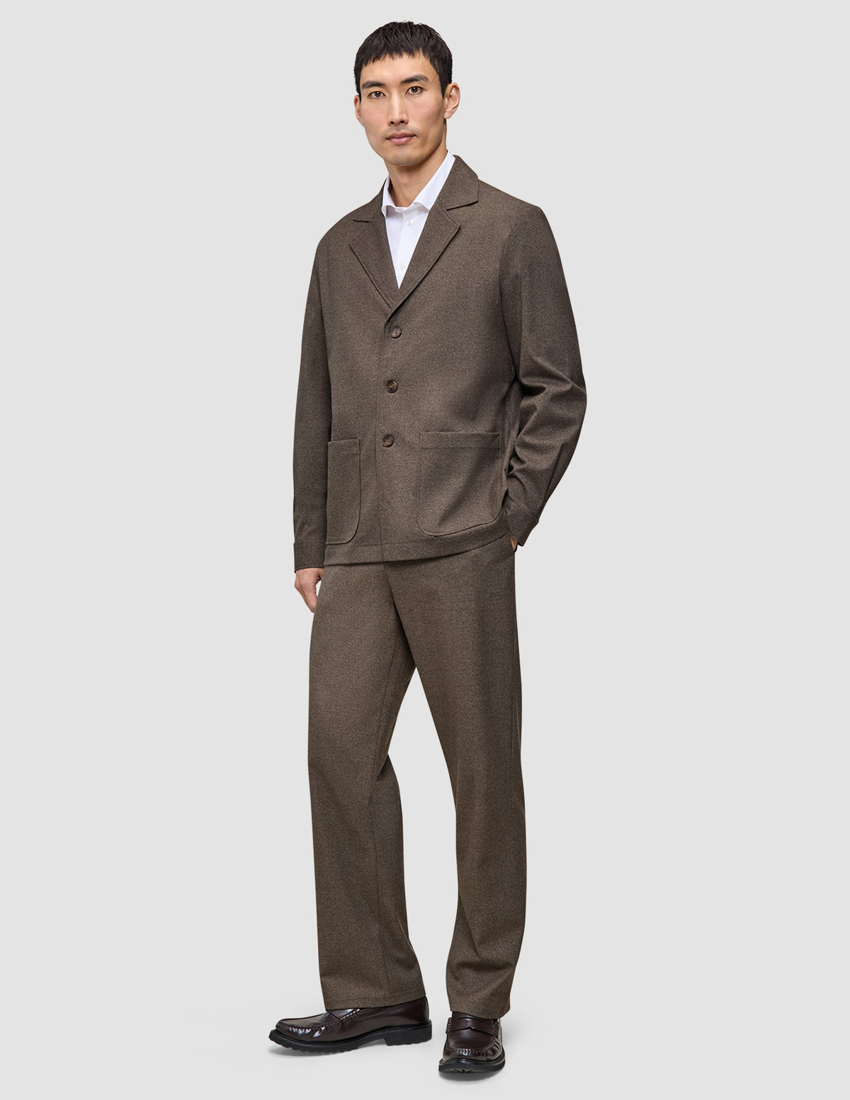 Ein Mann, der ein dunkelbeiges Atelier Overshirt aus gebrochenem Twill über einem weißen Hemd mit Kragen trägt, dazu eine passende Hose und dunkle Loafer im klaren, skandinavischen Design.