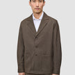 Ein Mann trägt ein Atelier Overshirt Dark Beige aus gebrochenem Twill mit Kragen, drei Knöpfen und zwei großen aufgesetzten Taschen über einem weißen Hemd mit Kragen, was ein klares, skandinavisches Design widerspiegelt.