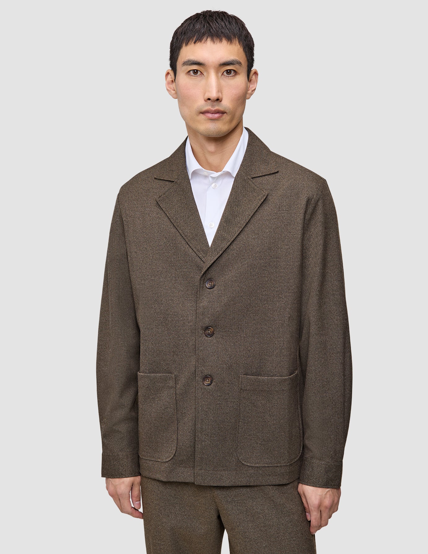 Ein Mann trägt ein Atelier Overshirt Dark Beige aus gebrochenem Twill mit Kragen, drei Knöpfen und zwei großen aufgesetzten Taschen über einem weißen Hemd mit Kragen, was ein klares, skandinavisches Design widerspiegelt.