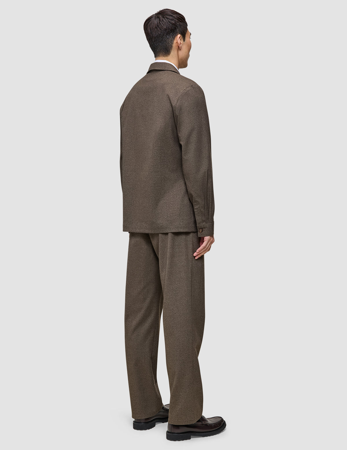 Ein Mann, von hinten gesehen, der ein Atelier Overshirt Dark Beige aus gebrochenem Twill trägt, dazu eine passende Hose und dunkle Loafer – ein Inbegriff des minimalistischen skandinavischen Designs.