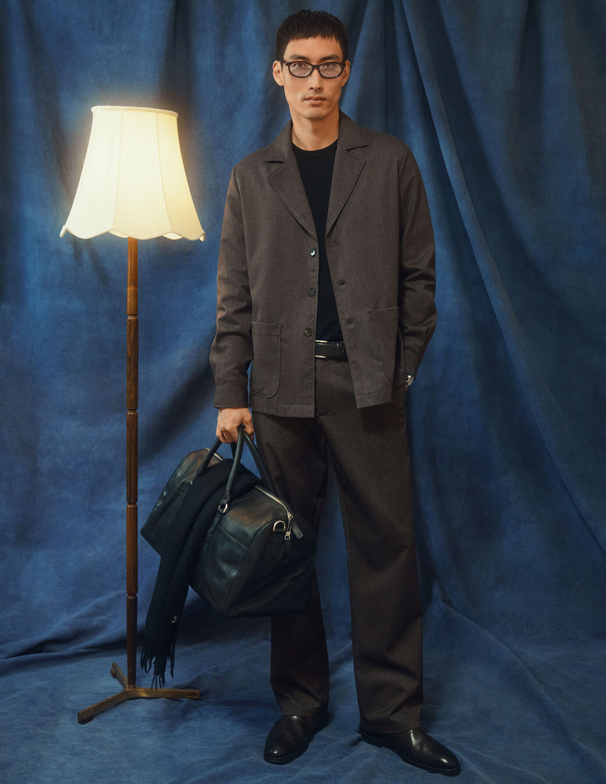 Eine Person, die ein dunkelbeiges Atelier Overshirt und eine dazu passende Hose aus gebrochenem Twill trägt, hält eine schwarze Reisetasche in der Hand.