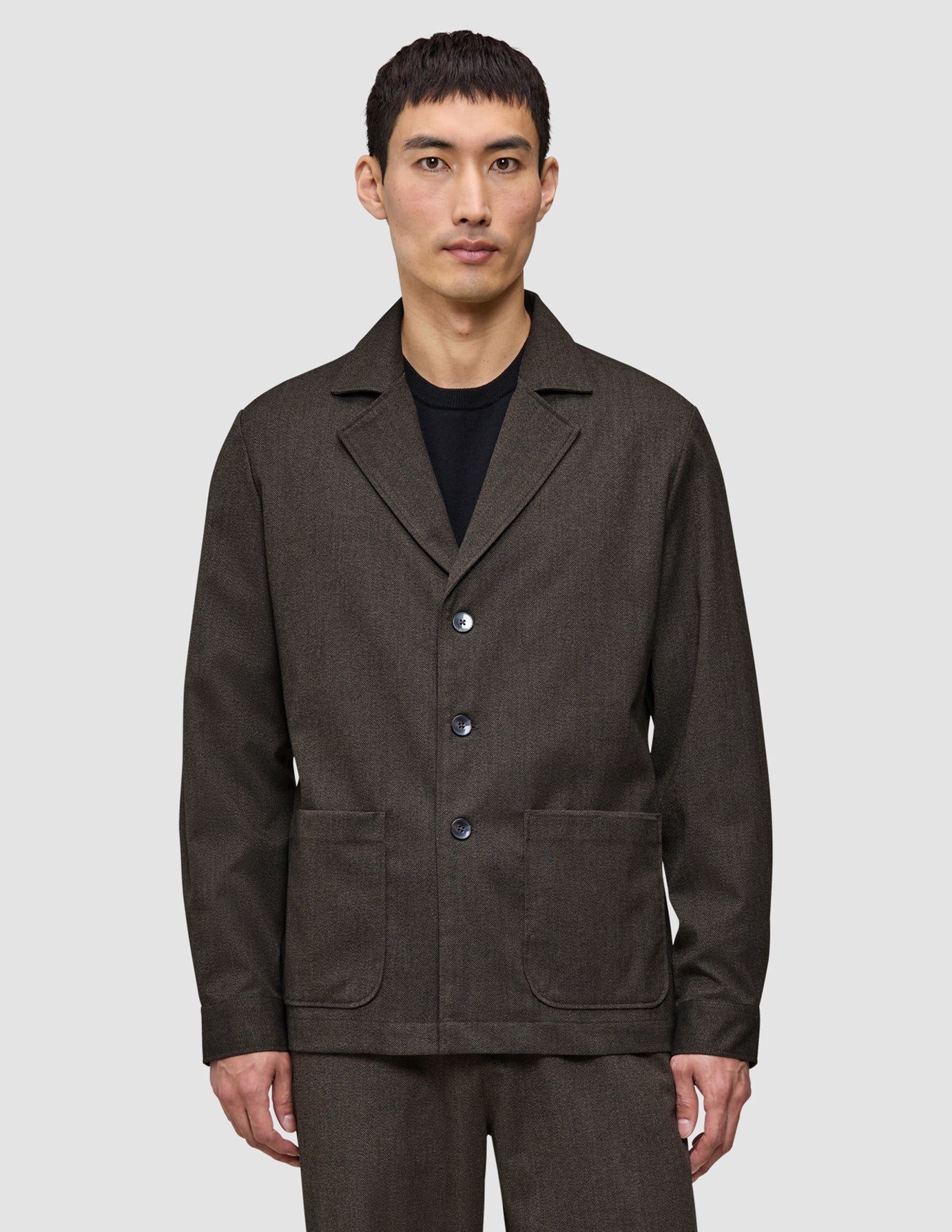 Ein Mann trägt ein dunkelbraunes Atelier-Overshirt aus gebrochenem Twill mit drei Knöpfen vorne und zwei aufgesetzten Taschen unten.