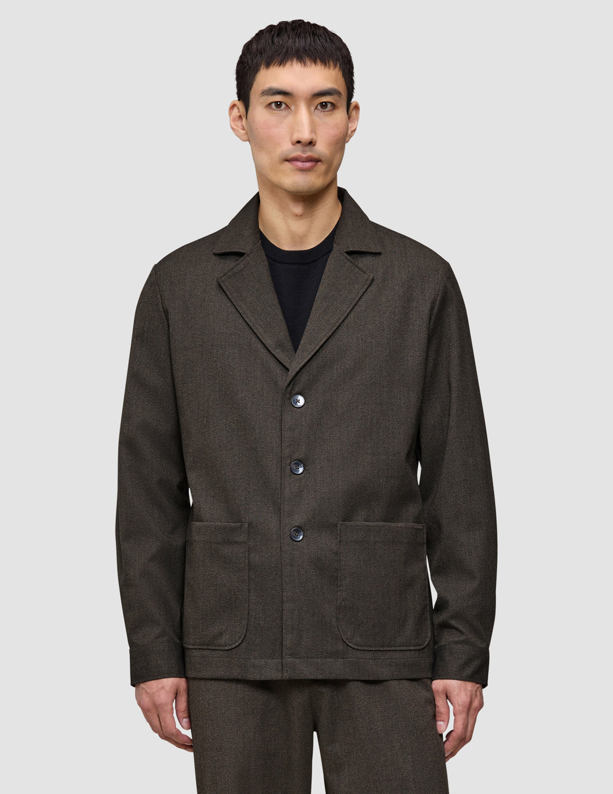 Ein Mann trägt ein dunkelbraunes Atelier-Overshirt aus gebrochenem Twill mit drei Knöpfen vorne und zwei aufgesetzten Taschen unten.