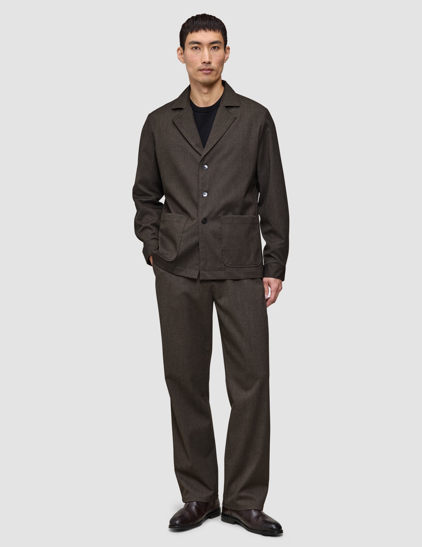 Ein Mann, der ein dunkelbraunes Atelier Overshirt aus gebrochenem Twill mit gekerbtem Kragen und einer dazu passenden Hose trägt.