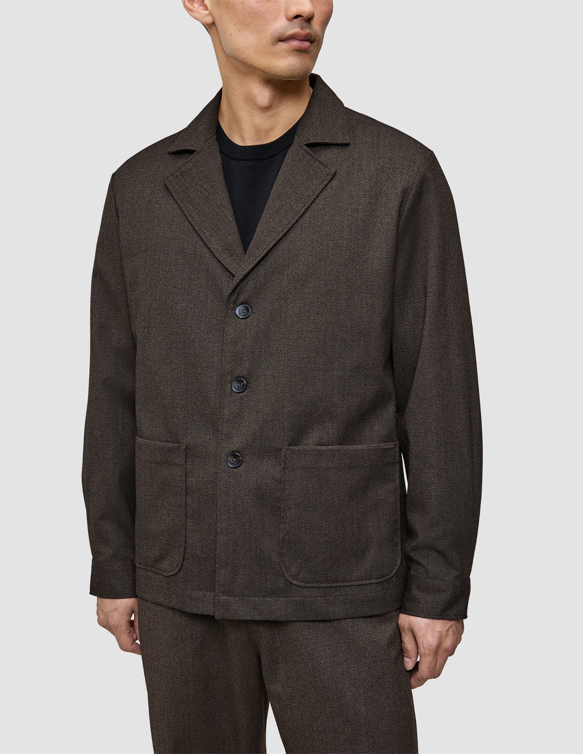 Ein Mann, der ein dunkelbraunes Atelier Overshirt aus gebrochenem Twill mit gekerbtem Kragen und zwei aufgesetzten Vordertaschen trägt.