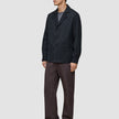 Ein Mann trägt ein dunkelblaues Atelier Overshirt über einem grauen T-Shirt, dazu eine Atelier-Hose und schwarze Loafer.