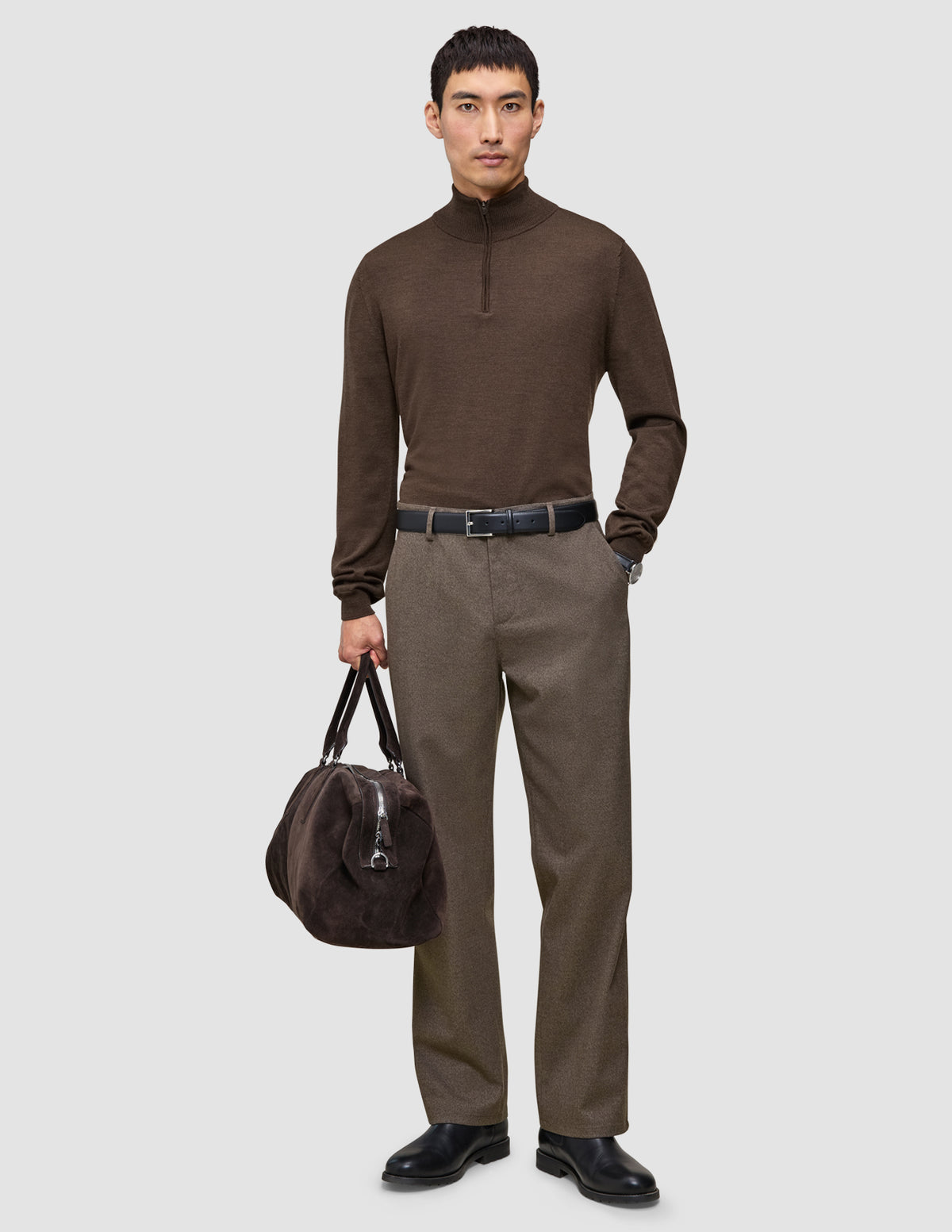 Ein Mann in einem braunen Pullover mit Viertelreißverschluss und einer Atelier Pants Relaxed in Dunkelbeige, der eine dunkelbraune Reisetasche im skandinavischen Design in der Hand hält.
