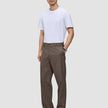 Ein Mann, der ein weißes T-Shirt und eine Atelier Pants Relaxed Dark Beige aus schwerem Broken-Twill trägt, dazu schwarze Lederschuhe.