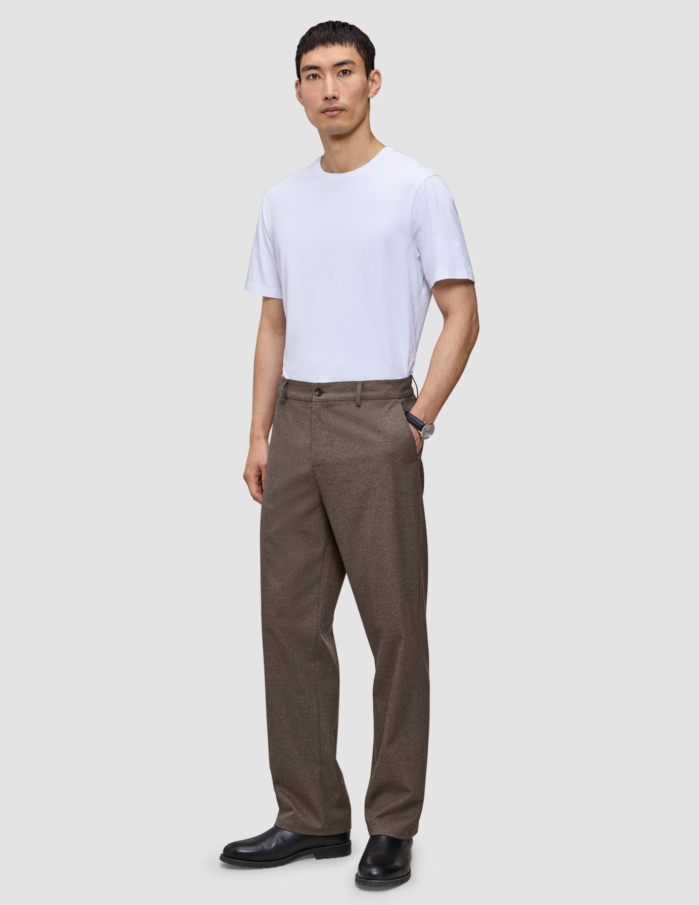 Ein Mann, der ein weißes T-Shirt und eine Atelier Pants Relaxed Dark Beige aus schwerem Broken-Twill trägt, dazu schwarze Lederschuhe.