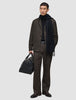 Ein Mann in einer dunkelbraunen Atelier Pants Relaxed, einem schwarzen Pullover und einem Schal, der eine schwarze Leder-Reisetasche in der Hand hält.
