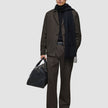 Ein Mann in einer dunkelbraunen Atelier Pants Relaxed, einem schwarzen Pullover und einem Schal, der eine schwarze Leder-Reisetasche in der Hand hält.