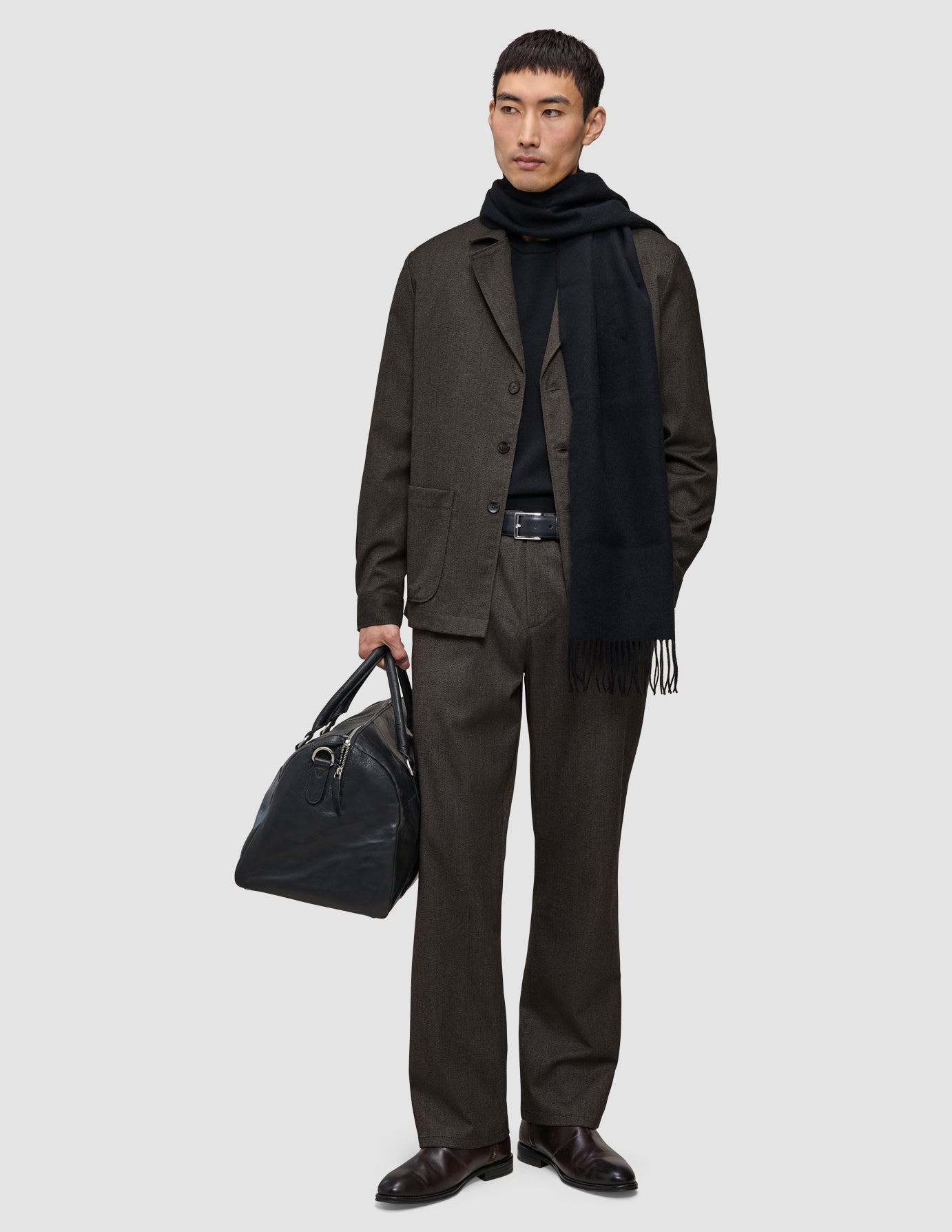 Ein Mann in einer dunkelbraunen Atelier Pants Relaxed, einem schwarzen Pullover und einem Schal, der eine schwarze Leder-Reisetasche in der Hand hält.
