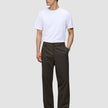 Ein Mann in einem weißen T-Shirt und einer Atelier Pants Relaxed in Dunkelbraun aus schwerem, gebrochenem Twill, die das minimalistische skandinavische Design verkörpert.