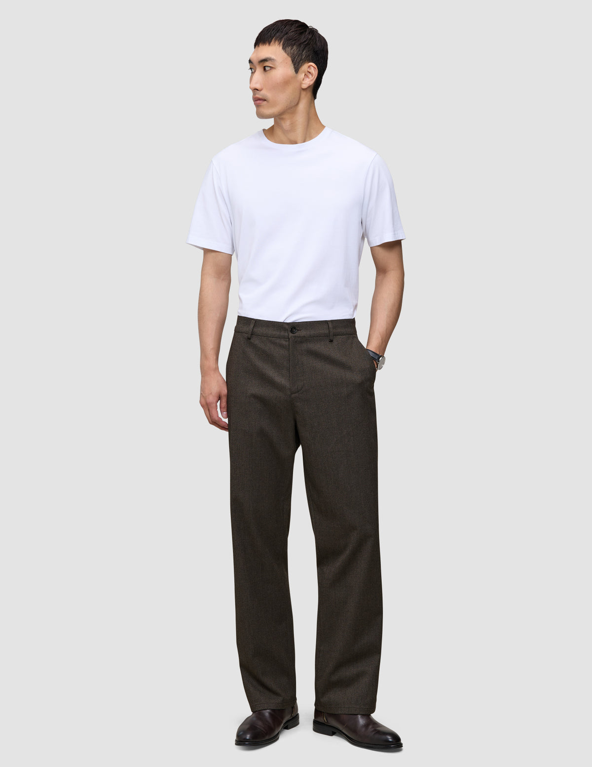 Ein Mann in einem weißen T-Shirt und einer Atelier Pants Relaxed in Dunkelbraun aus schwerem, gebrochenem Twill, die das minimalistische skandinavische Design verkörpert.